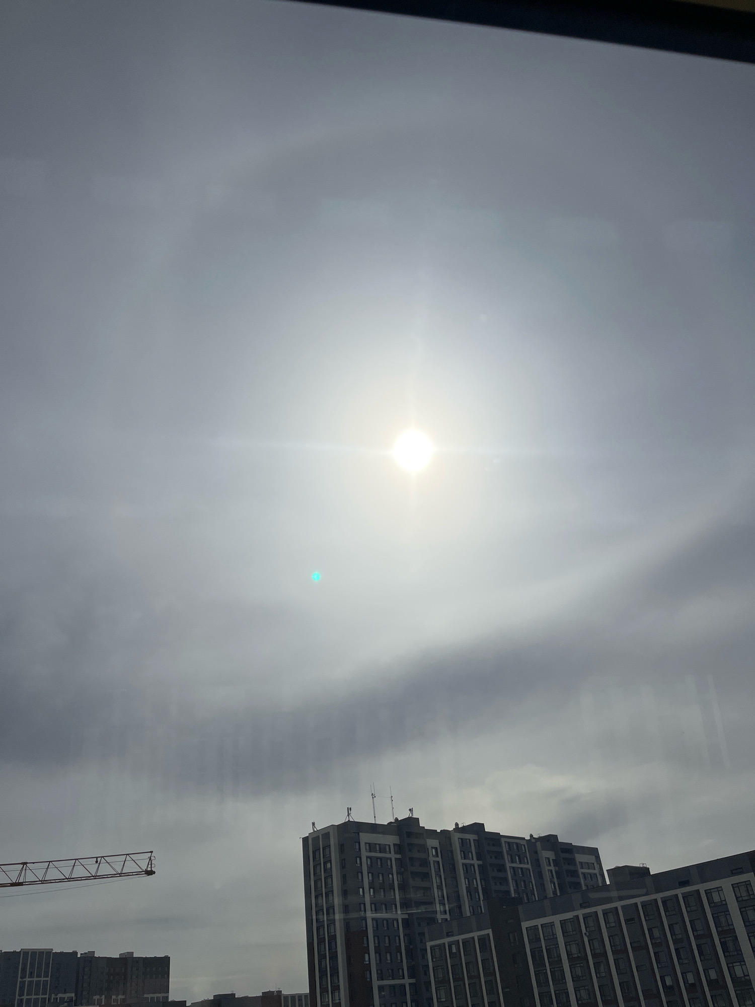 The image depicts an overcast sky with the sun faintly visible as a bright, diffuse light source near the center. A faint solar halo is observable, a ring of light around the sun. In the lower portion of the image, a construction crane extends from the left, and several modern multi-story buildings are visible in the right and lower portions of the frame. The scene appears to be an urban environment. The specific location within Astana, Kazakhstan, cannot be determined from the visual information alone. There are no people or discernible actions in the image.