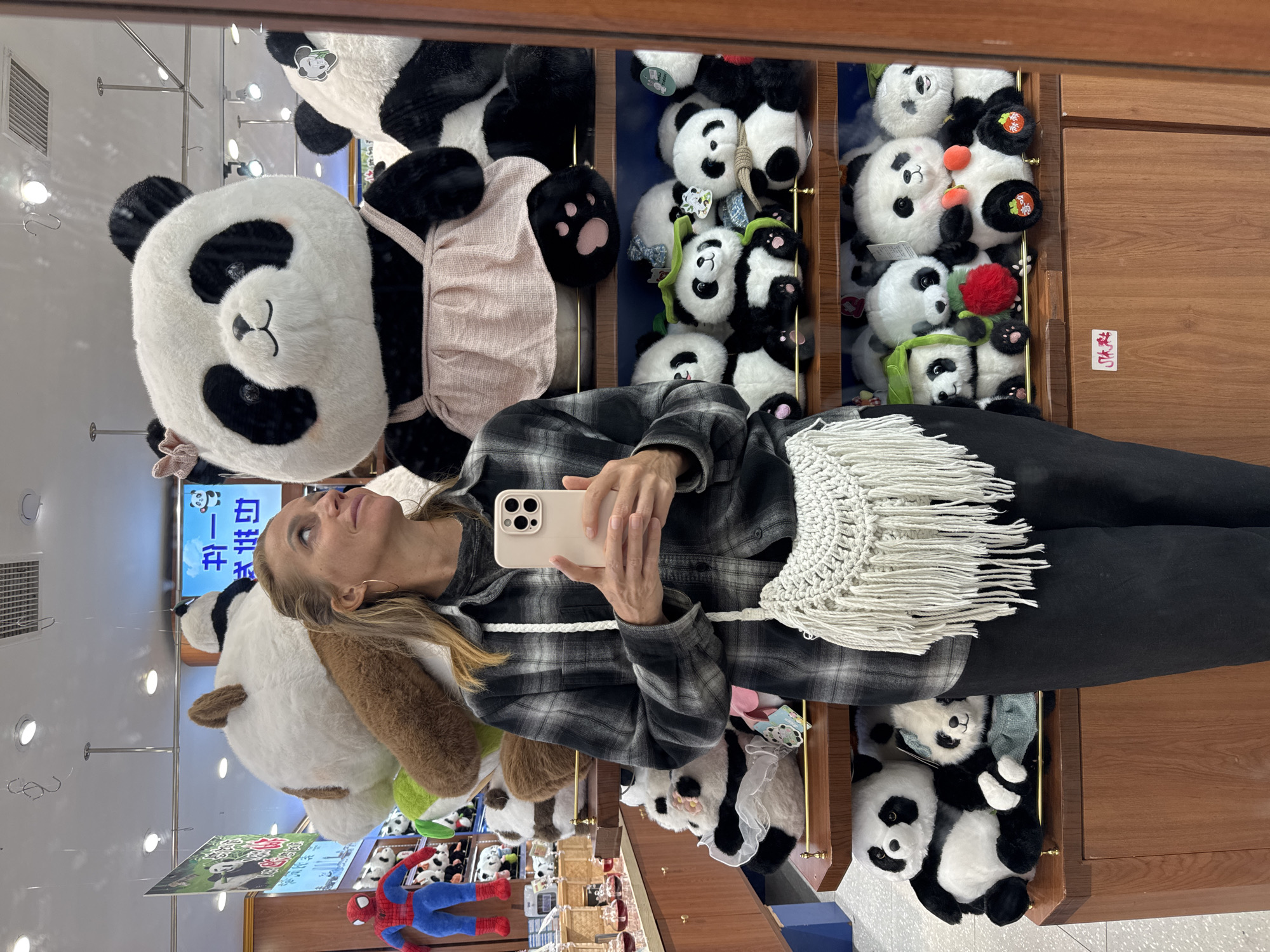 In Jinjiang District, China, a woman with fair skin, blonde hair, and wearing a black and white plaid shirt and black pants holds a light-colored smartphone in her hands and takes a mirror selfie in a shop selling panda plush toys. Behind the woman, the shelves are filled with various panda plushies, arranged in rows. The environment includes light-colored walls, shelves with plush pandas, and other merchandise visible in the periphery. The lighting is bright and artificial, typical of a retail environment. The focus is on the woman and the numerous panda toys behind her.