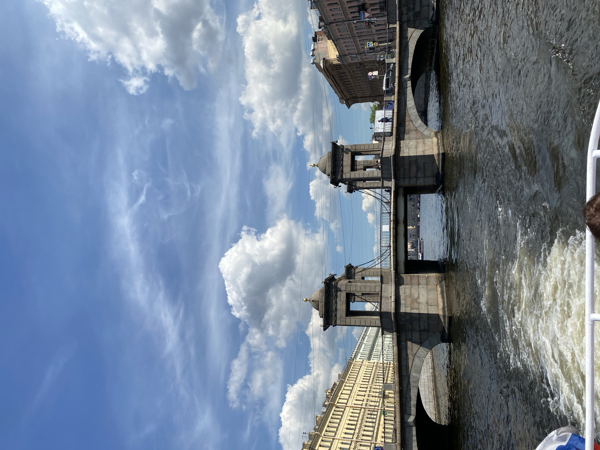 The image shows a view of the Anichkov Bridge over the Fontanka River in Saint Petersburg, Russia. The bridge is a stone arch bridge with two large stone piers. The piers are topped with decorative domes. The bridge is lined with buildings on either side of the river. The sky is blue with some white clouds. The water in the river is dark and choppy, with a wake from a boat in the foreground. A small part of a boat with a Russian flag is visible at the bottom left of the frame. There are a few people visible on the bridge and in the distance. The buildings appear to be historic, with multiple stories and ornate facades.