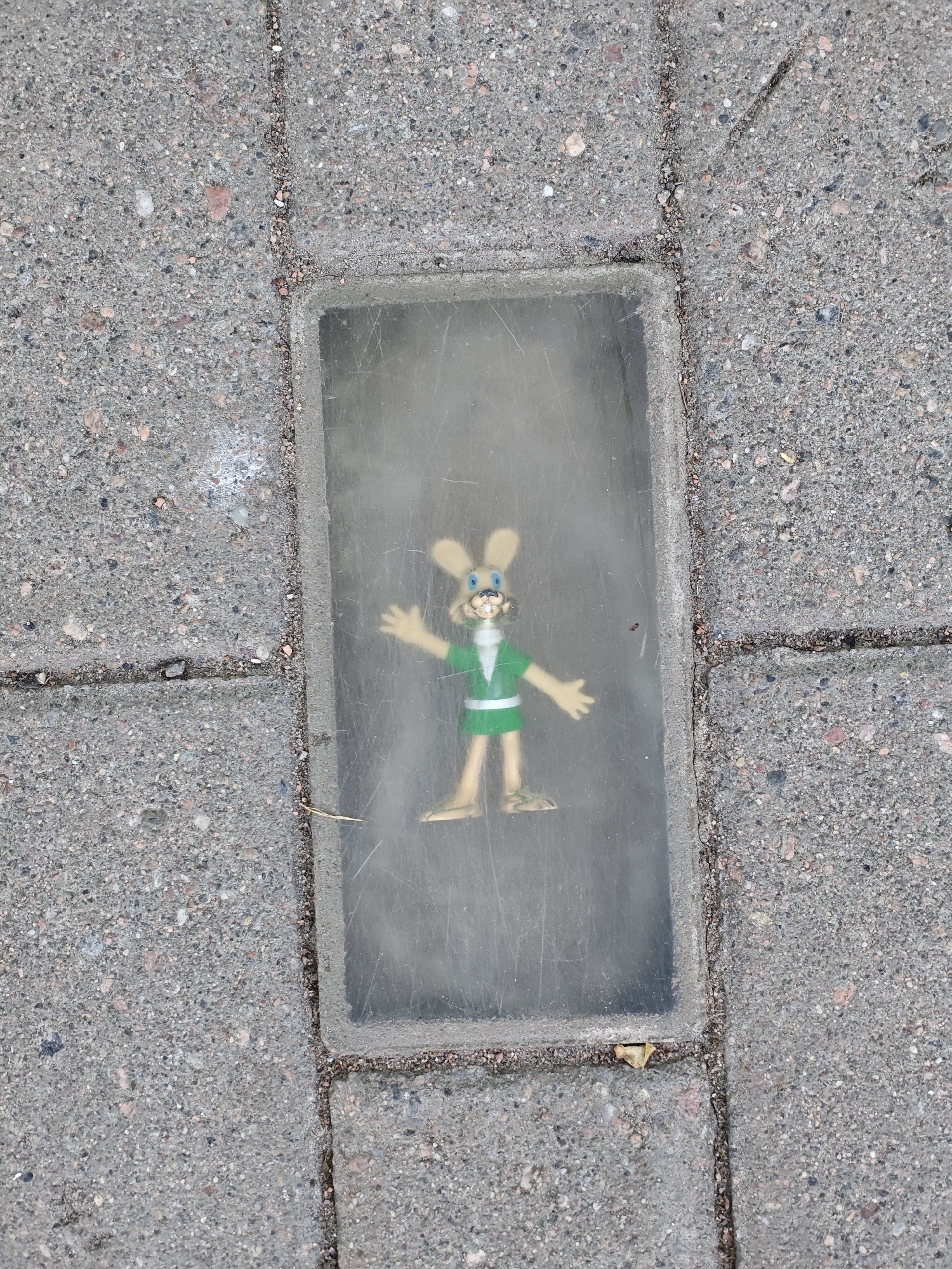A plastic figurine of a cartoon rabbit character stands inside a rectangular, recessed glass or plastic casing embedded in a sidewalk. The rabbit is light brown with long ears, blue eyes, and a wide smile. It wears a green shirt with a white collar and waistband, and tan shorts. Its arms are raised slightly outwards, as if waving. The casing appears slightly dusty or smudged, obscuring a clear view of the figurine. The sidewalk is made of grey concrete pavers interspersed with small pebbles and gravel. The scene is captured from a top-down perspective. The lighting suggests it is daytime, likely outdoors, given the natural light illuminating the scene and the texture of the concrete. No discernible text is visible in the image. The location context is Saint Petersburg, Russia, though no specific landmarks or cues confirm this within the image itself.