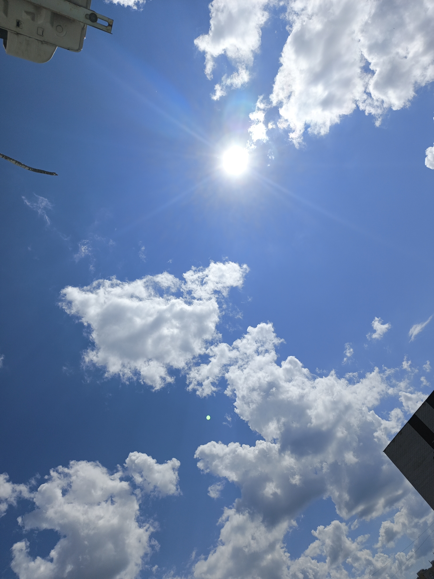 The image captures a bright, sunlit sky with scattered white cumulus clouds. The sun is prominently visible, emitting bright rays. In the upper left quadrant, a portion of a light-colored, industrial-looking structure is visible, possibly a part of a building or equipment. A thin, dark, linear object extends diagonally across the sky from the left edge. In the lower right, a dark, angled surface with what appears to be a reflective pattern or text is partially visible. The overall scene is an outdoor, daytime environment with a clear blue sky. The location is identified as Kazan, Russia, but there are no specific landmarks or contextual clues within the image to confirm this. No people or animals are present.
