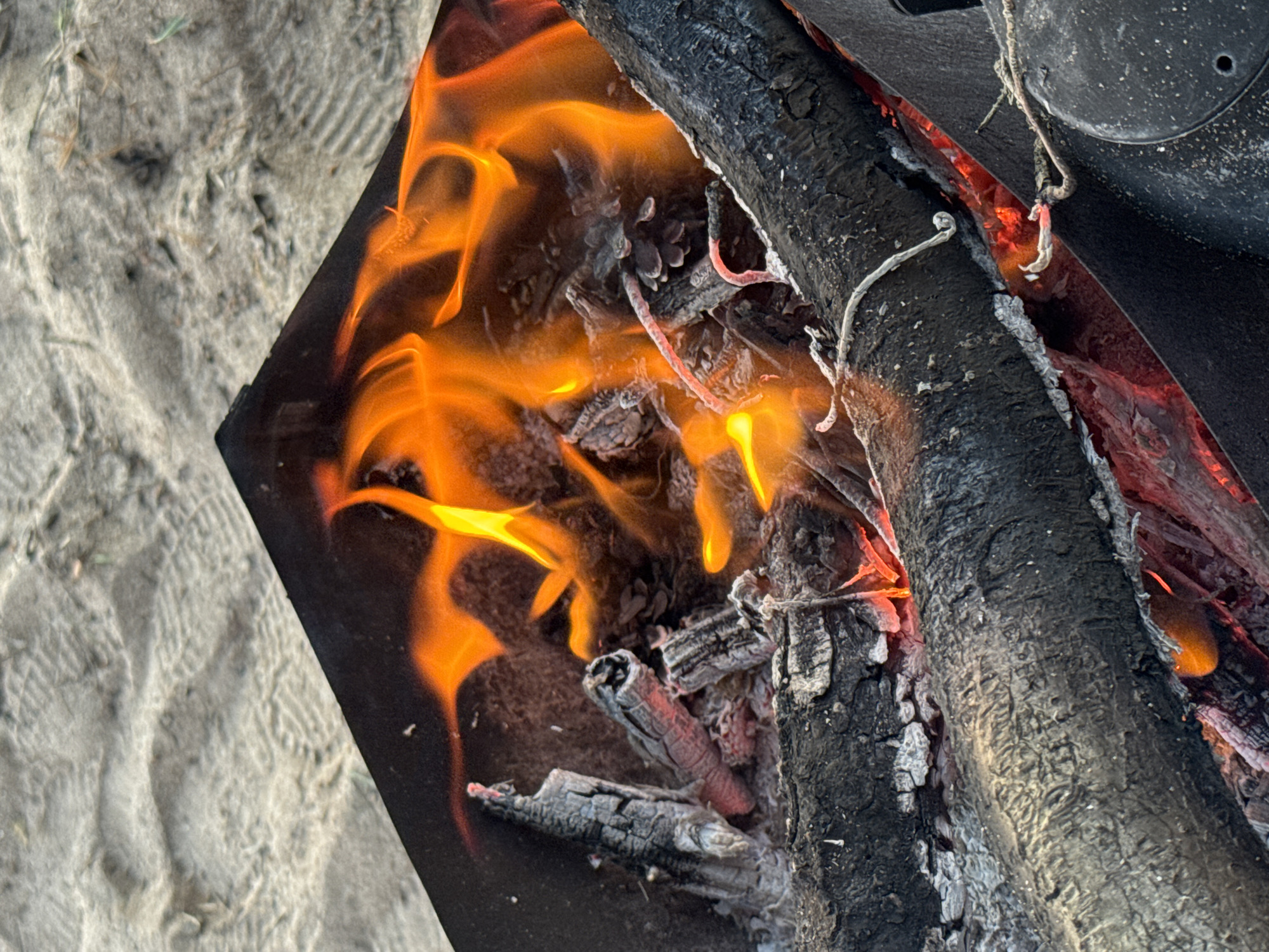 In Shepelevo, Russia, an outdoor scene depicts a close-up view of a campfire burning in a black metal fire pit, situated on sandy ground. The fire, characterized by vibrant orange flames and glowing embers, consumes logs, pinecones, and ash. The surrounding sand shows faint imprints, possibly from footwear, suggesting human presence in the area.