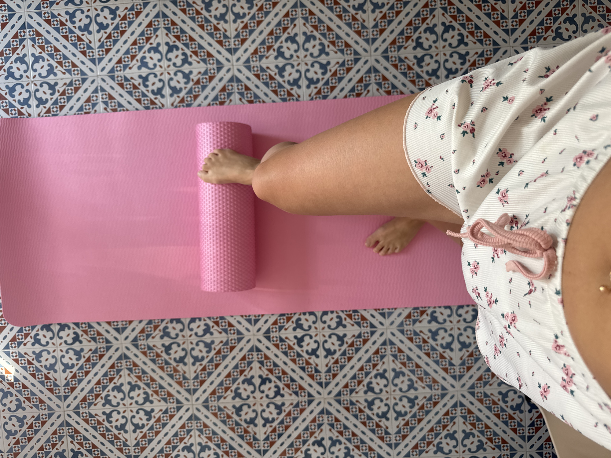 The image shows a person's legs and abdomen on a pink yoga mat. The yoga mat is placed on a floor with a patterned tile design. A pink foam roller is positioned on the yoga mat, with the person's right foot resting on it. The person is wearing white shorts with a floral print. The scene appears to be indoors, possibly in a home or gym setting. The presence of the yoga mat and foam roller suggests an activity related to fitness or recovery. The location of Isola delle Femmine, Italy is not discernible from the visual information provided in the image.