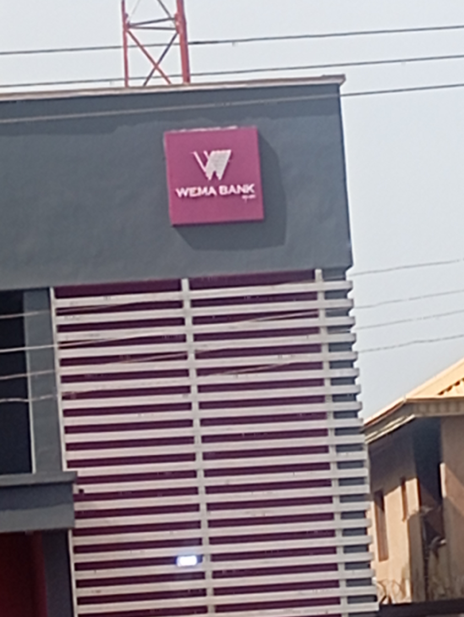 The image shows the exterior of a building with a sign that reads "WEMA BANK". The building has a modern design with a dark gray facade and a section with horizontal purple and white louvers. To the right of the building, a portion of another structure with a light-colored roof and windows is visible. The sky is overcast. A red metal tower-like structure is partially visible in the upper background. The scene appears to be located in an urban or semi-urban area.