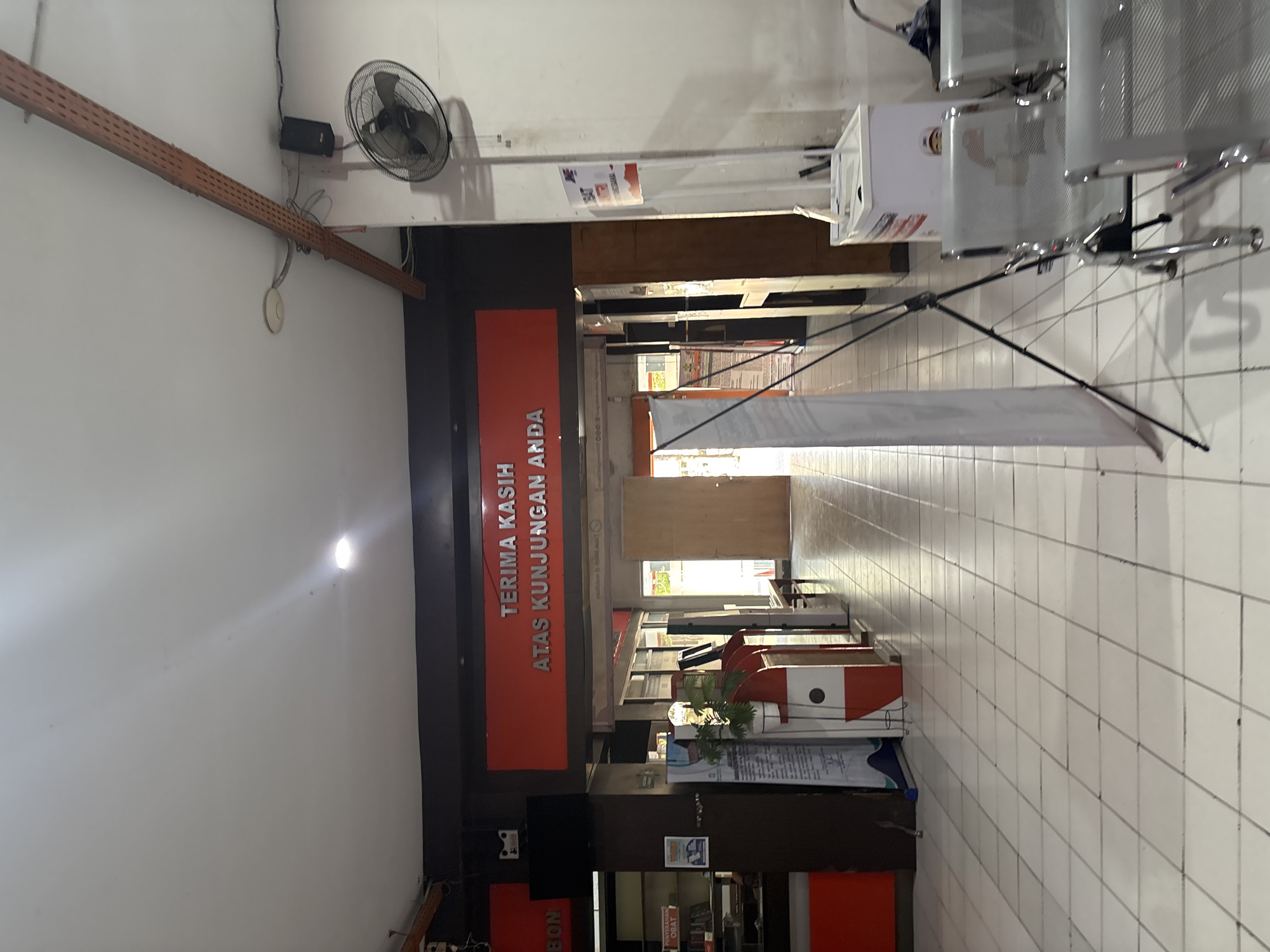 The image depicts the interior of a waiting area, likely a public facility in Cirebon, Indonesia, given the Indonesian text on the signage. Dominating the frame is a large, red sign with white lettering that reads "TERIMA KASIH ATAS KUNJUNGAN ANDA," which translates to "THANK YOU FOR YOUR VISIT." The room features a white tiled floor and a white tiled ceiling with recessed lighting.
To the left, there is a dark brown cabinet with a television mounted on top. Below the television, what appears to be a display or counter is visible. Further to the left, red accents are present on what might be a counter or furniture. A white standing banner with text and graphics is also visible on the left side of the hallway.
In the center of the hallway, a closed wooden door is prominent. To the right of the door, a series of silver chairs with metallic frames are arranged in rows, suggesting a waiting area. A tall, white stand with a black fan is mounted on the wall to the right. Another white refrigerator-like unit with graphics is positioned near the chairs. A black tripod stands near the chairs, holding a white banner or screen.
The environment appears to be clean and well-lit, with a sense of order. The overall impression is that of a service area or waiting room within a government office, post office, or similar establishment. The lighting suggests it could be daytime, with artificial lights supplementing natural light from an unseen source. No individuals are clearly visible in the frame.