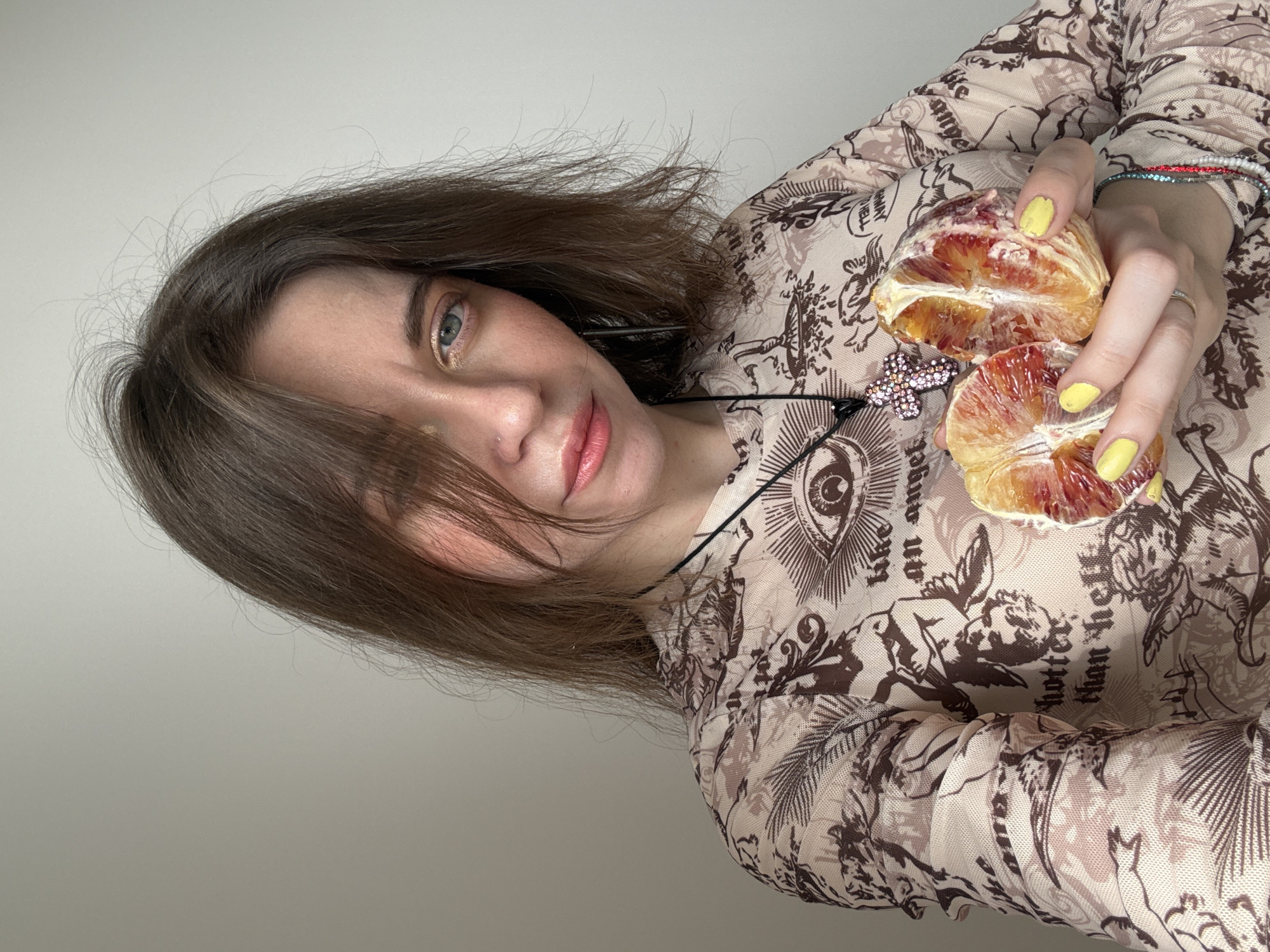 A young woman with short brown hair is holding two halves of a blood orange. She is wearing a patterned top with images of angels and eyes. Her fingernails are painted yellow. The location is difficult to determine from the image, as it lacks contextual elements that point to Moscow, Russia.