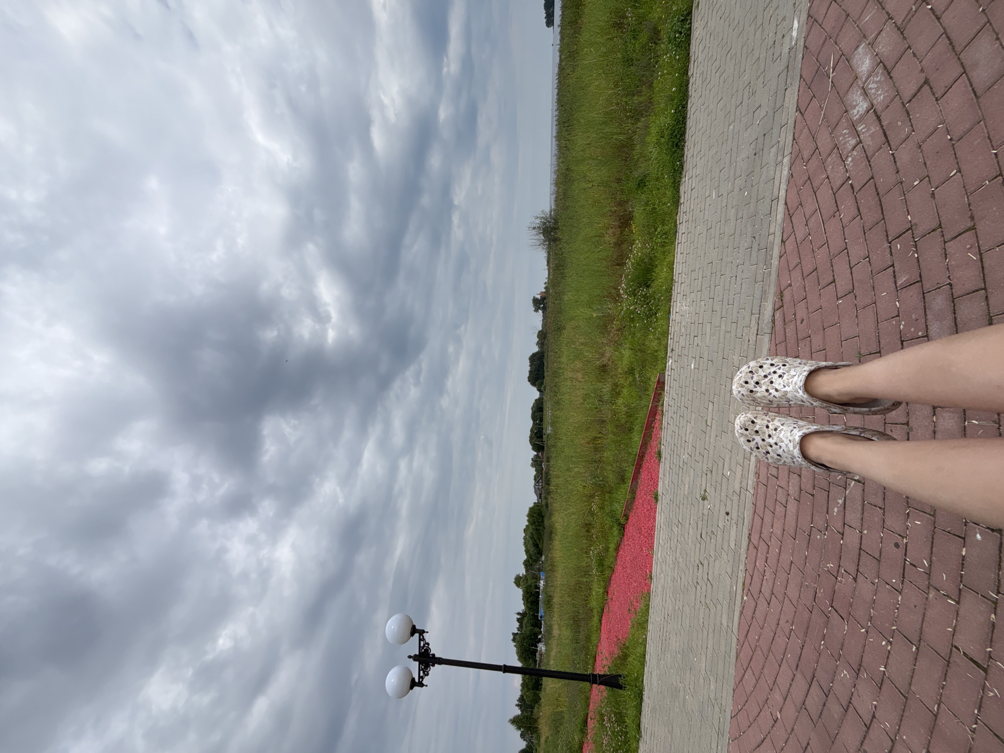 In Breytovo, Russia, a first-person perspective shot captures a person's legs, adorned with white patterned clogs, resting on a curved brick-paved area. The pathway is edged with red gravel and green grass. A dual-headed lamppost stands to the left, its white globes illuminated against a cloudy, overcast sky. Beyond the greenery, a body of water is visible, with a distant treeline and scattered buildings on the horizon.