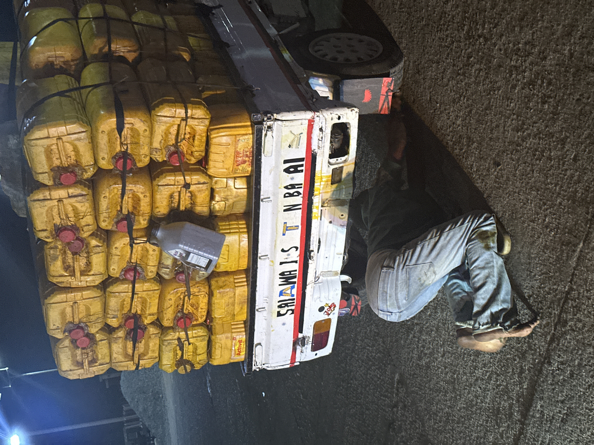 The image depicts a white truck loaded with yellow jerrycans secured with black straps. The truck has the name "SALAWATS NBA BAI" written on its side in blue and red letters. A man wearing blue jeans and a dark shirt is lying on the ground near the truck, and he appears to be barefoot. The setting appears to be an outdoor location, and the location is specified as Gura Top, Nigeria.