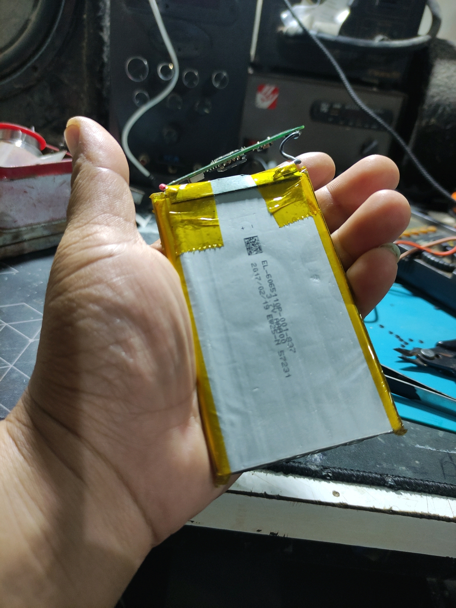 A person's hand is holding a flat, rectangular battery with yellow tape along its edges. A small green circuit board is attached to one end of the battery, with a thin wire connecting to it. The background is cluttered with electronic equipment, tools, and a black workbench surface with a blue mat. The scene appears to be a workshop or repair area, but its specific location within Bulukumba, Indonesia cannot be determined from the image alone.