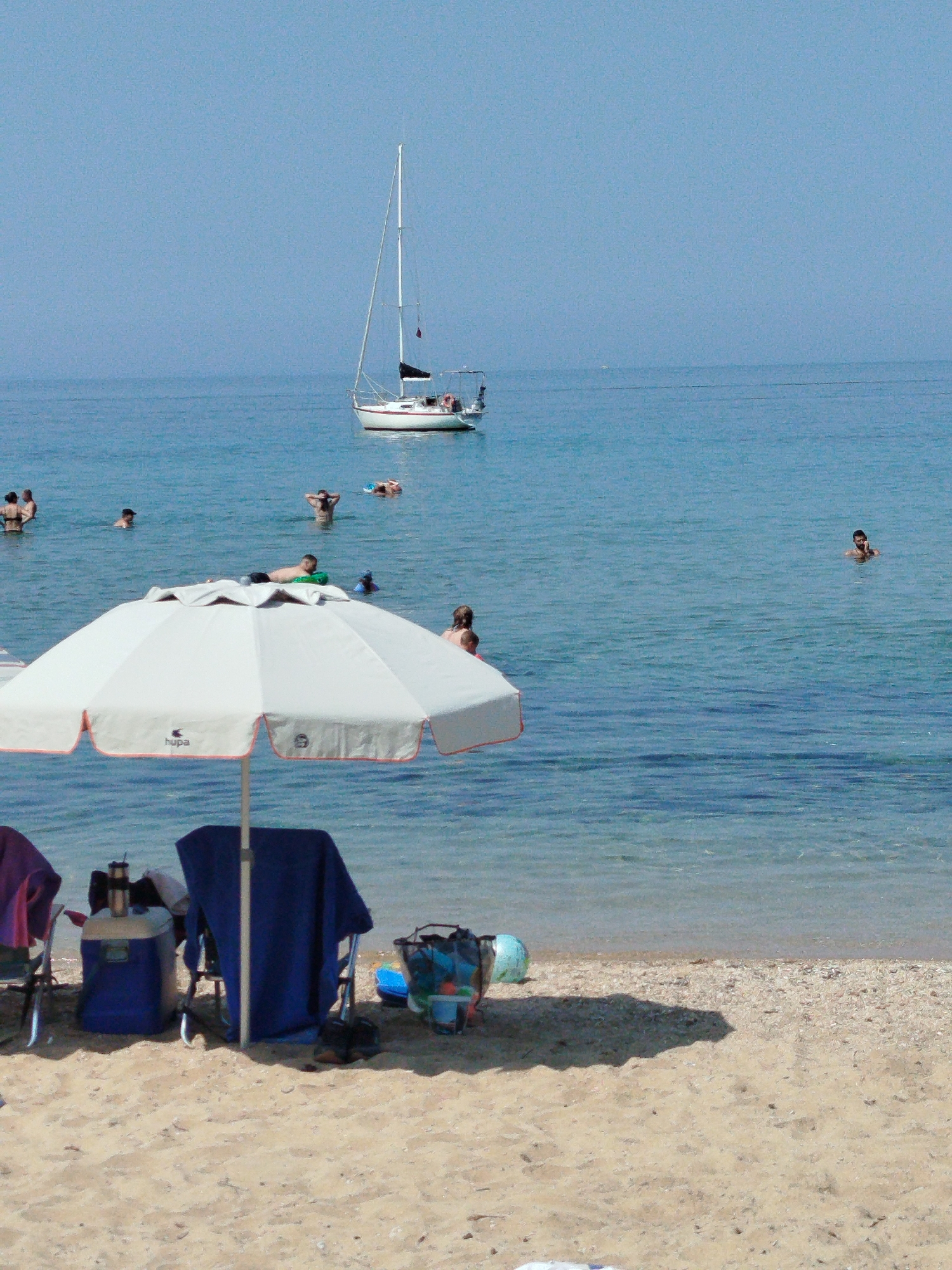 The image depicts a sunny day at a beach in Sithonia Municipal Unit, Greece. In the foreground, a white beach umbrella with orange trim is set up on the sandy shore, casting a shadow. To the left of the umbrella are two chairs, one with a dark purple towel draped over it, and a blue cooler with a thermos. To the right of the umbrella, a blue towel is draped over a chair. Further to the right, a black mesh bag filled with beach items and a blue inflatable ball are visible.
The sea is calm and clear blue, with several people enjoying a swim. In the middle ground, a white sailboat with a black sail is anchored. In the background, the horizon is a clear blue sky, merging with the sea. The overall scene is one of a relaxed beach day.