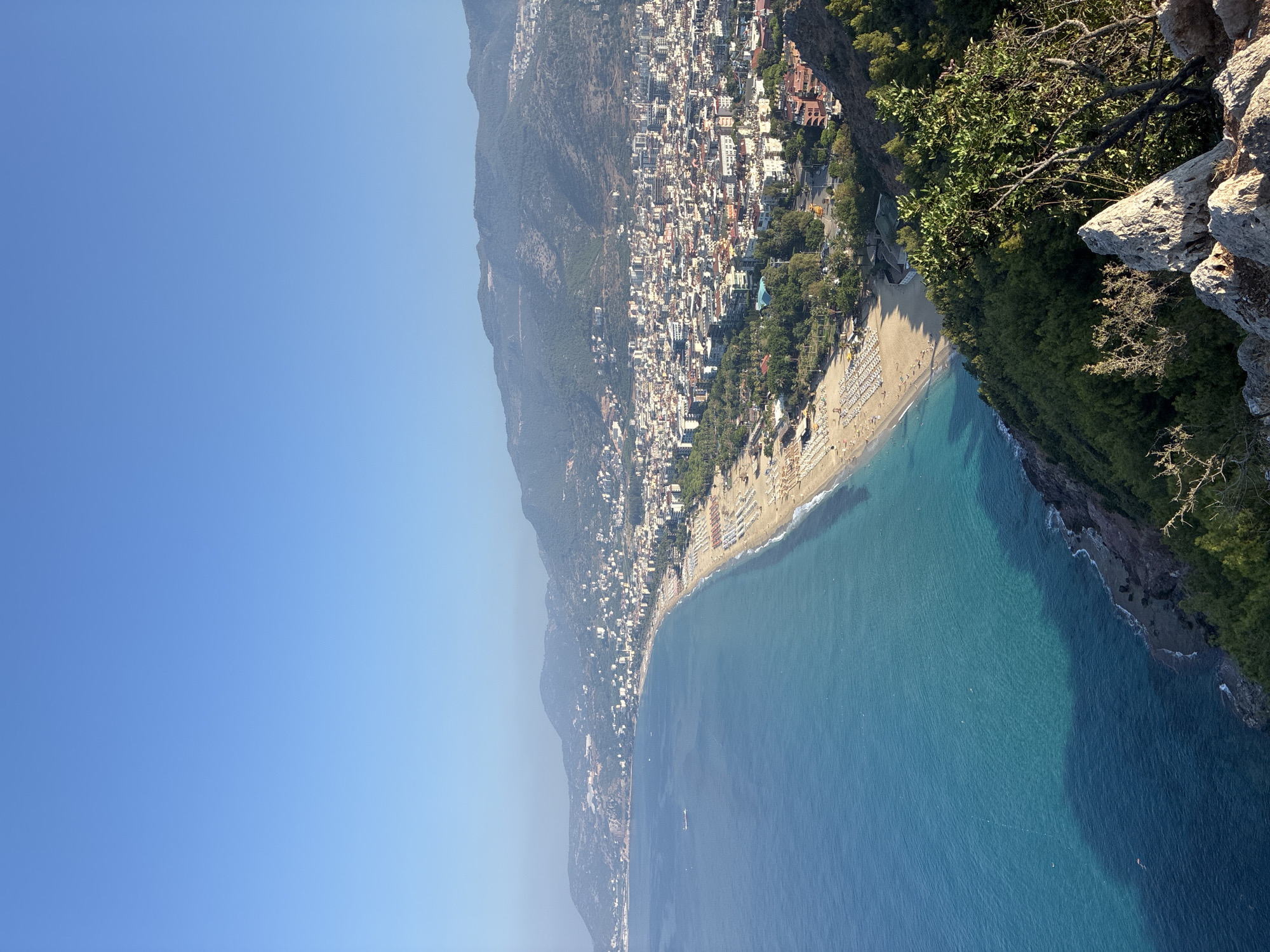 The image captures a panoramic view of Alanya, Turkey, from an elevated vantage point. The scene overlooks a coastline characterized by a crescent-shaped beach adorned with numerous rows of white lounge chairs and umbrellas. The turquoise waters of the sea gently lap against the sandy shore, with a distinct contrast in water depth indicated by a darker blue shadow stretching across the sea.
Beyond the beach, a densely populated city unfolds, with buildings of varying heights and architectural styles nestled against a backdrop of rolling, tree-covered hills and mountains that extend into the hazy distance. The urban landscape features a mix of white, cream, and terracotta-colored buildings, interspersed with patches of greenery. A road with some visible traffic runs along the edge of the beach.
In the foreground, weathered rocks and lush green foliage are visible, framing the descent towards the city and the sea. The sky is a clear, unblemished blue, suggesting a bright, sunny day. A single white boat is observed in the distance on the water. The overall impression is one of a vibrant coastal city, with its beach, urban sprawl, and natural mountainous environment coexisting under a clear sky.