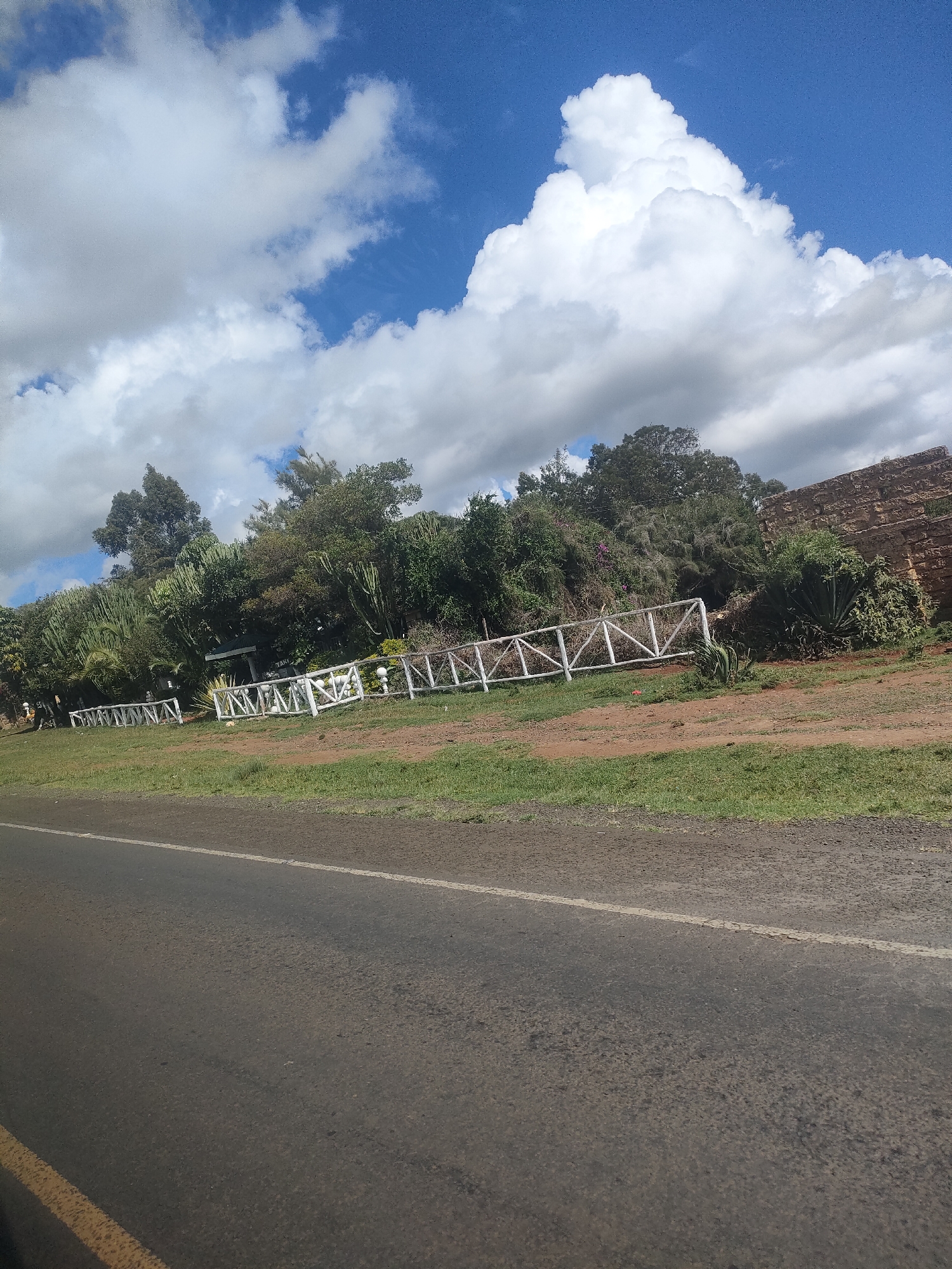 The image shows a section of a road in Kisima ward, Kenya under a partly cloudy sky. The road is asphalt with a faded white line marking. Adjacent to the road is a strip of grass, followed by a dirt area bordered by a white wooden fence. Behind the fence is dense vegetation, including trees and cacti. A section of a stone wall is visible in the background. No people or animals are visible in the immediate scene.
