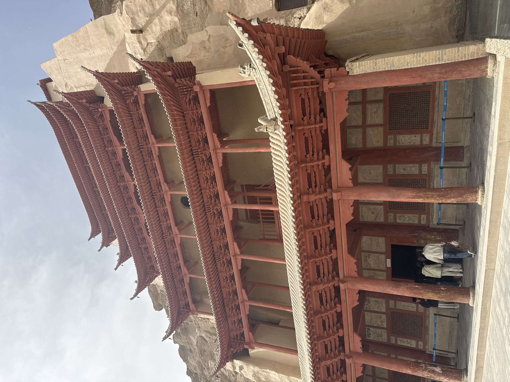 The image shows the exterior of a traditional Chinese building structure in Dunhuang City, China. The building has multiple tiers of red-painted wooden beams and supports, with intricately carved details. The roof has projecting eaves. People are visible near the entrance. The structure appears to be built against a rock cliff face. The sky is visible in the background.
