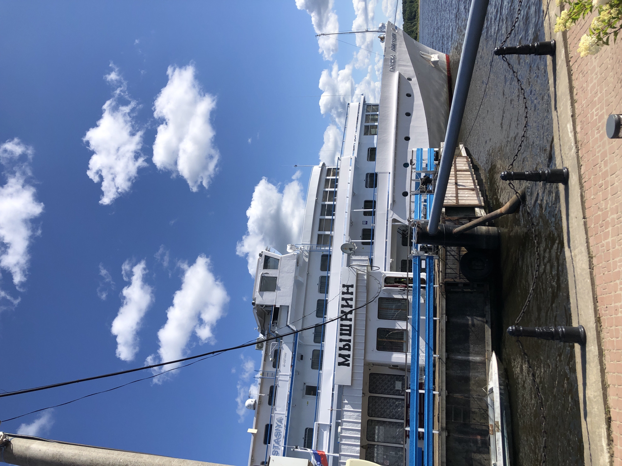 A white passenger ship, the "Myshkin," is docked at a pier in Myshkin, Russia, on a sunny day with a blue sky and scattered white clouds. The ship is large and multi-decked, with numerous windows and a blue railing along its lower deck. A small white boat is tied alongside the ship. The pier is made of brick and concrete, with black metal bollards and chains lining the edge. The water of the Volga River is visible, reflecting the sky and the ship. Trees line the far bank of the river.