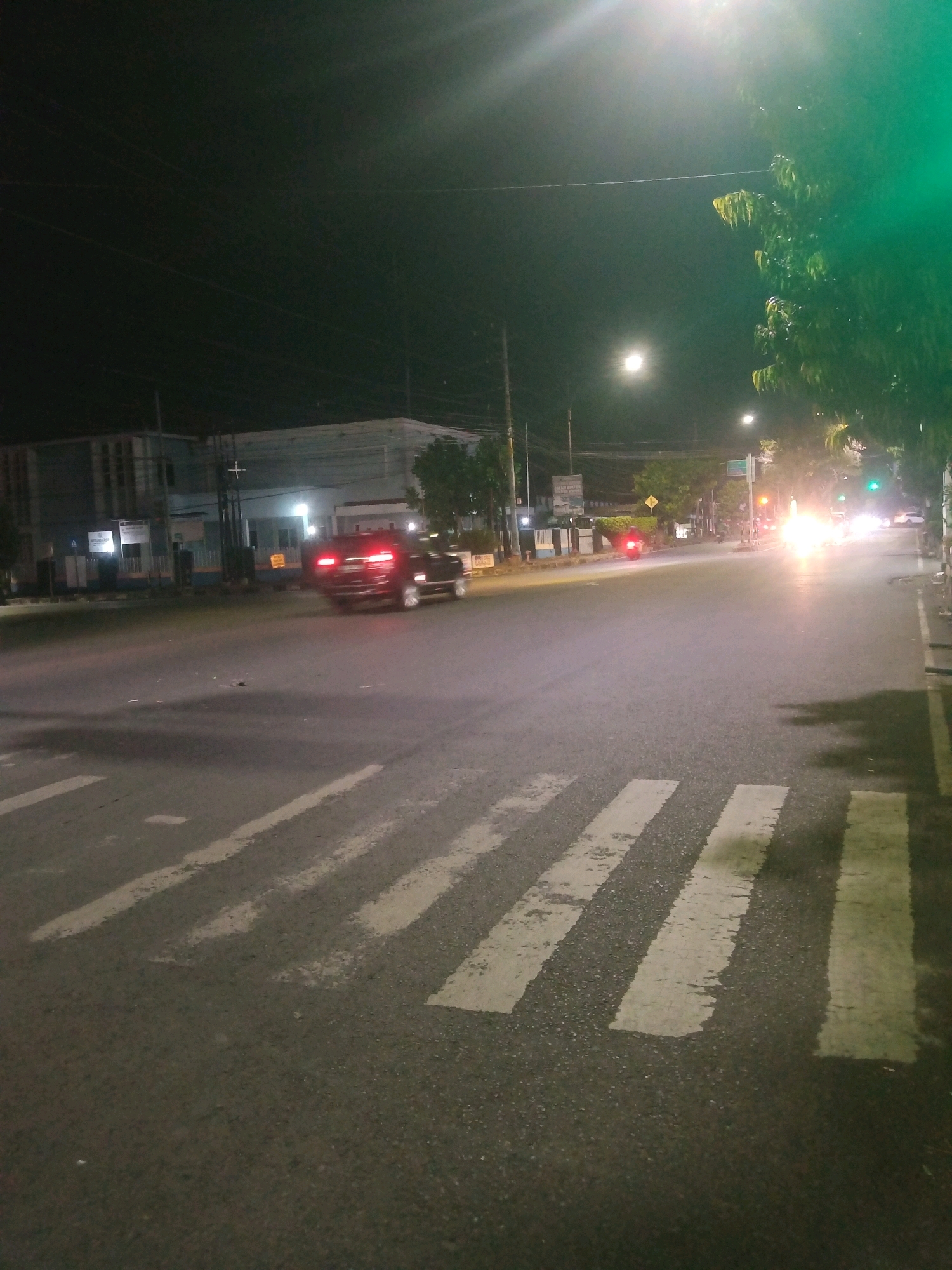 A nighttime, eye-level, wide shot captures a crosswalk on a dimly lit, wet asphalt road in Purwokerto, Indonesia. In the foreground, several white stripes of the crosswalk are visible. A dark-colored car is in motion in the middle ground, its brake lights illuminated. To the left of the road, buildings line the street, some with illuminated windows. Trees and streetlights are scattered throughout the scene, with a bright light source emanating from the upper right quadrant of the image, casting a green hue. The sky is dark, suggesting it is late at night.