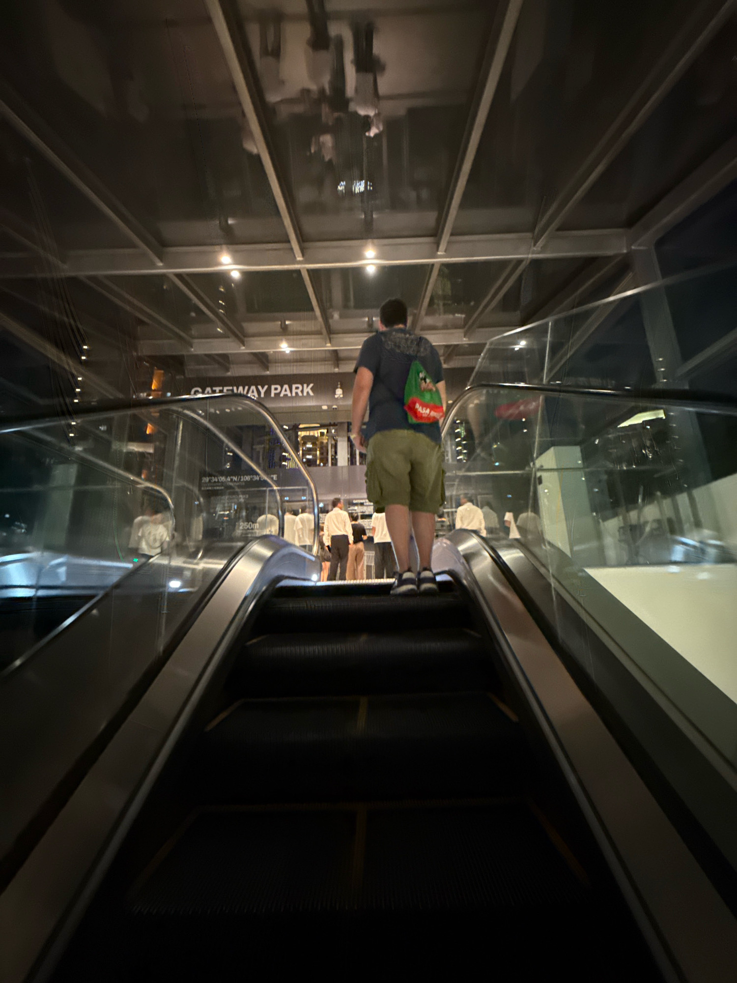 In Yuzhong District, China, a person is riding up an escalator. The individual, seen from behind, is wearing a dark t-shirt, olive-green cargo shorts, and sneakers. A colorful tote bag is slung over their left shoulder. The escalator has dark treads with yellow safety lines. The scene is indoors, under a ceiling with a glass and metal structure. In the background, a sign reads "GATEWAY PARK." Other individuals are visible further down the escalator and on the level above. The lighting suggests it is nighttime or an interior space.