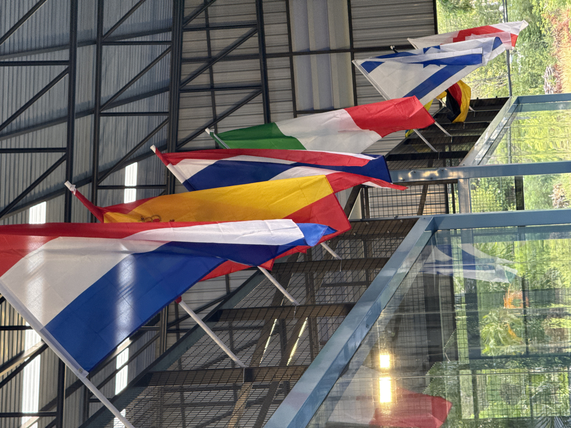 The image shows a series of national flags hanging from the ceiling of an indoor structure, possibly a covered sports facility, in Ko Samui District, Thailand. The flags are displayed in a line, with some overlapping. Visible flags include the Netherlands, Spain, Russia, Italy, and Israel. A portion of the German flag is also visible. The structure has a metal roof and supporting beams, and a wire mesh fence runs along a mezzanine level. In the foreground, a reflective surface, likely glass, shows reflections of the surrounding environment, including greenery and artificial lighting. There are no people visible in the image. The overall scene suggests an event or establishment that celebrates international diversity, such as a sports complex or a cultural venue.