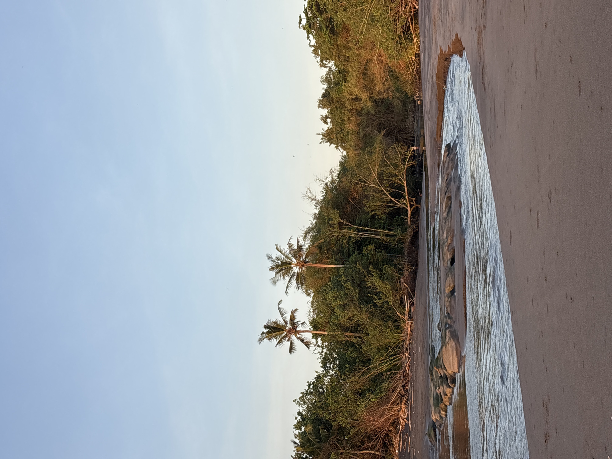 The photograph shows a beach scene in Kerambitan, Indonesia. A stream of water runs vertically between a dark sand beach and dense vegetation, including palm trees. The sky is light blue.