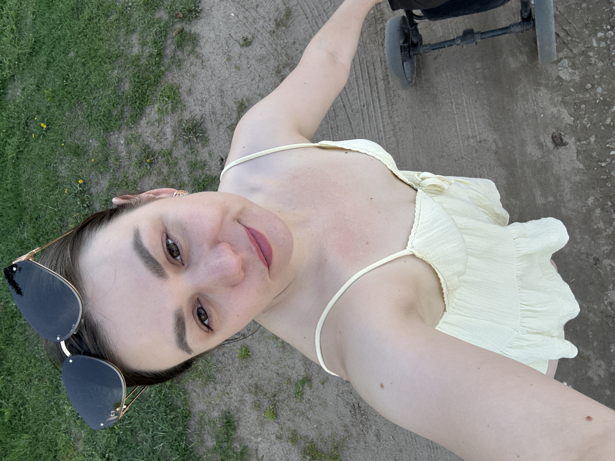 A young woman with sunglasses is taking a selfie while pushing a stroller on a dirt path. The path is bordered by grass on one side. The location is possibly in Chaikovsky, Russia, based on the prompt.