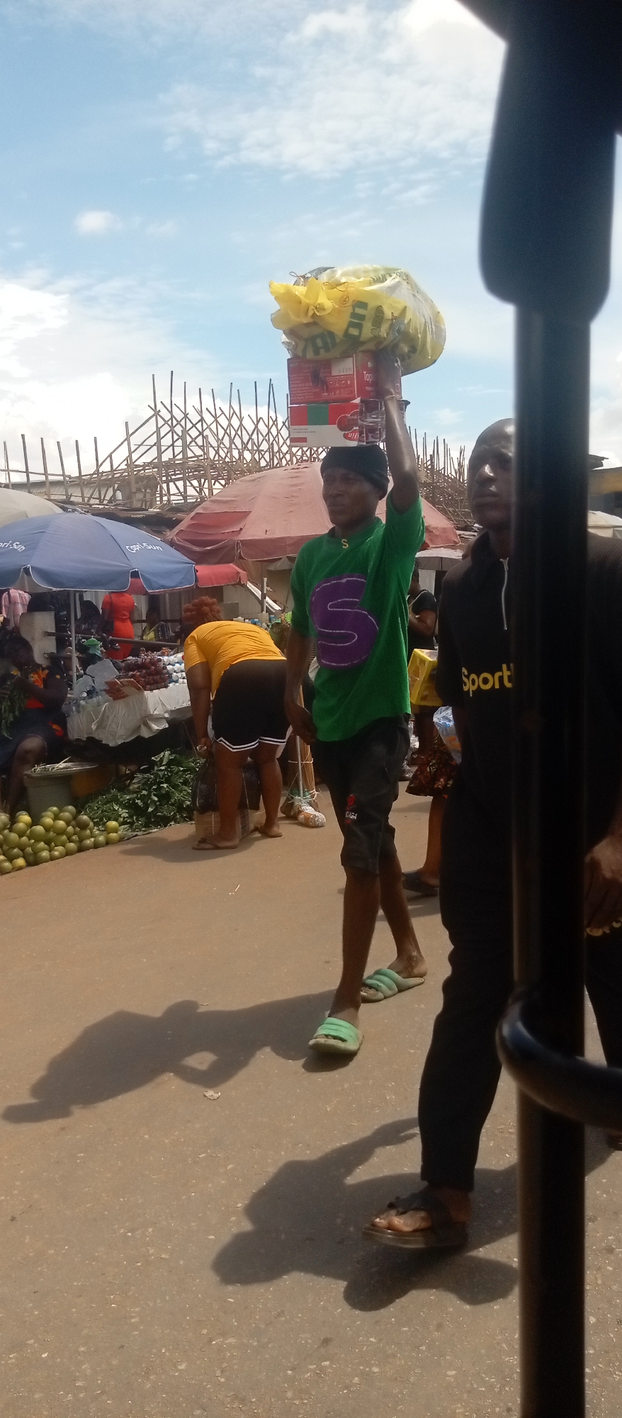 The image shows a bustling street scene, likely a marketplace, in the city of Unknown, Unknown. A person wearing a green shirt with a purple "S" is carrying a load of items balanced on their head. To their left is a vendor in a yellow shirt. Further in the background, there are market stalls with blue and red umbrellas. A wooden structure is visible overhead. To the right of the person carrying the load, another person wearing a black shirt that says "Sport" in yellow letters is partially visible. The lighting is bright, casting strong shadows on the ground.