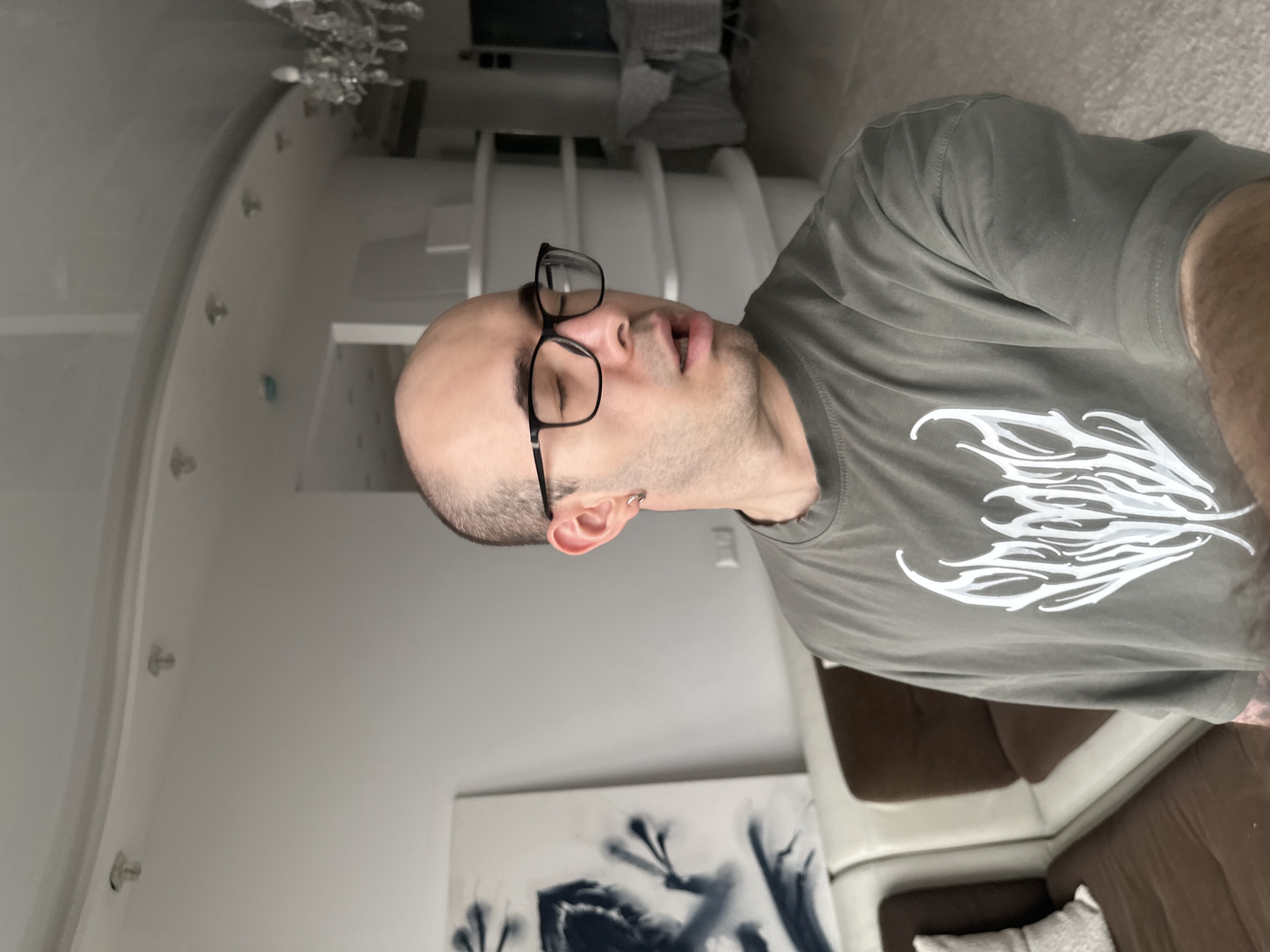 A man with a shaved head, wearing glasses and an olive green t-shirt with a white design, is lying down with his eyes closed and mouth slightly open. He has a silver earring in his left ear. Behind him is a white wall with a curved design and a decorative chandelier. In the lower part of the frame is an abstract black and white painting and a brown and white sectional couch. It is not possible to verify that the location is in Khimki, Russia based on the provided image.