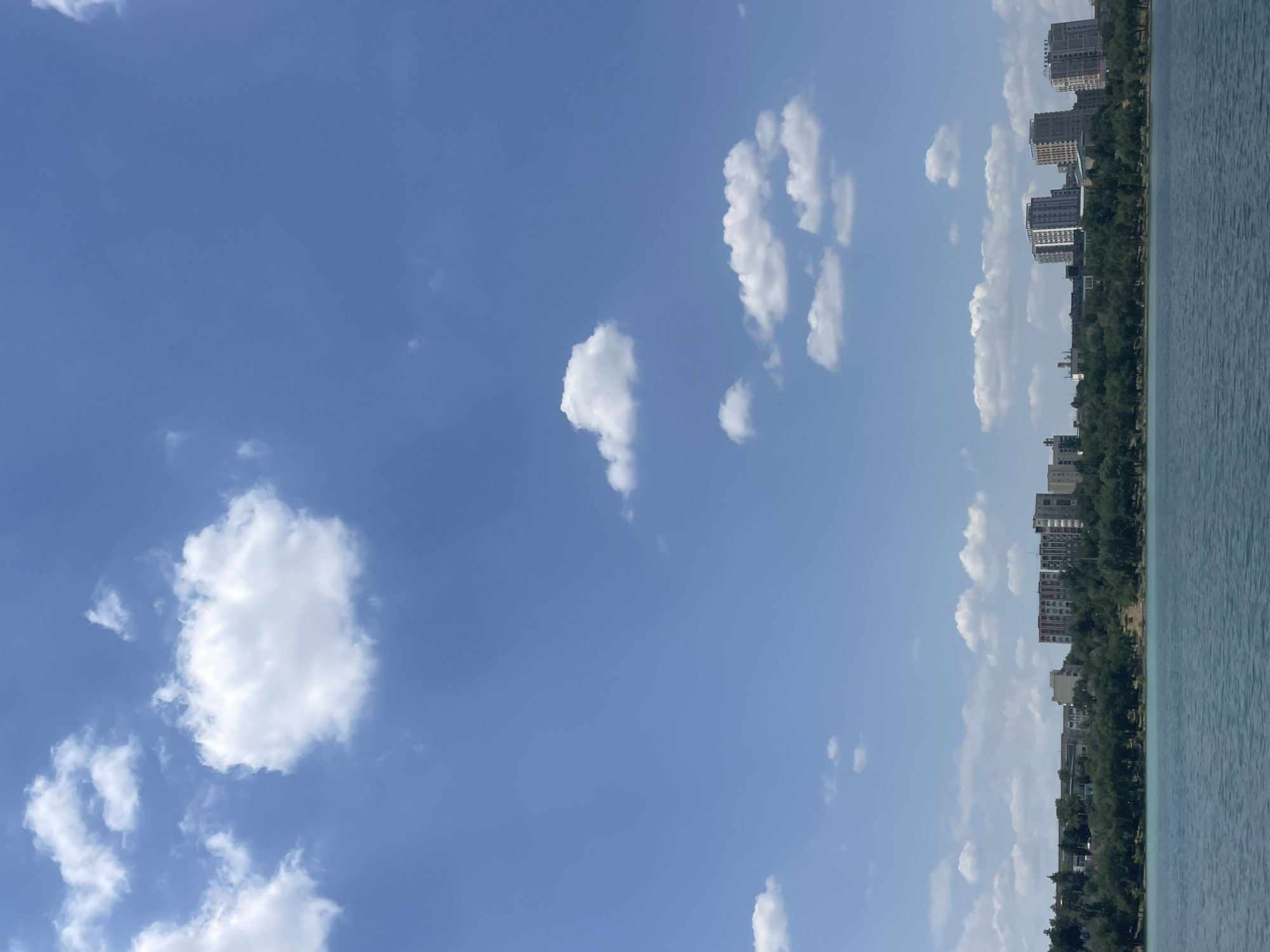 This is a horizontal, full-shot, eye-level photograph capturing a serene waterscape in Almaty, Kazakhstan. The foreground is dominated by a large body of calm, blue-green water, likely a lake or reservoir, with gentle ripples disturbing its surface. Beyond the water, a dense line of lush green trees forms a natural border along the shoreline. Behind the trees, a cityscape unfolds, characterized by several multi-story buildings of varying architectural styles and heights. Some of these buildings are residential apartments, appearing modern and well-maintained, while others could be commercial or administrative structures. The buildings are situated on a gentle slope, with the tallest ones visible in the middle and right sections of the frame.
The sky above is a clear, vibrant blue, punctuated by scattered white, fluffy cumulus clouds. The sunlight appears bright, suggesting it is daytime, likely late morning or afternoon, with the sun not directly visible. The overall atmosphere is peaceful and tranquil, showcasing a harmonious blend of nature and urban development. The location is identifiable as Almaty, Kazakhstan, specifically the area around the Almaty Lake, or Sary-Arka Lake, known for its scenic beauty and recreational opportunities.