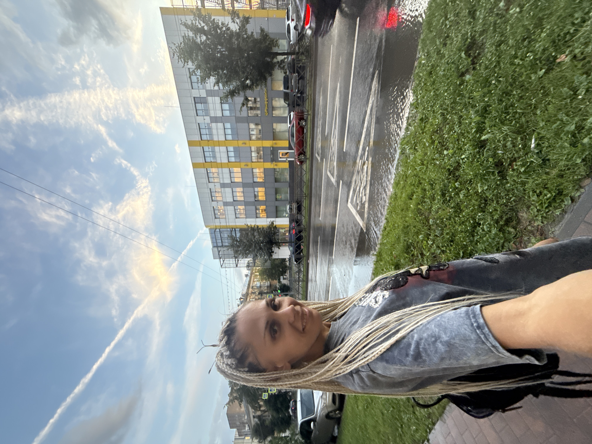 The image is a selfie taken by a woman with blonde braided hair, standing on a sidewalk with green grass next to a wet street. She is wearing a grey t-shirt and a backpack. Behind her, across the street, is a modern, multi-story building with a grey and yellow facade. The street appears wet, suggesting recent rain, with reflections of lights visible on the surface. There are cars parked and driving on the street, as well as a few trees. The sky above is blue with white clouds, and a contrail from an aircraft is visible. The scene is urban, with elements of a city environment.