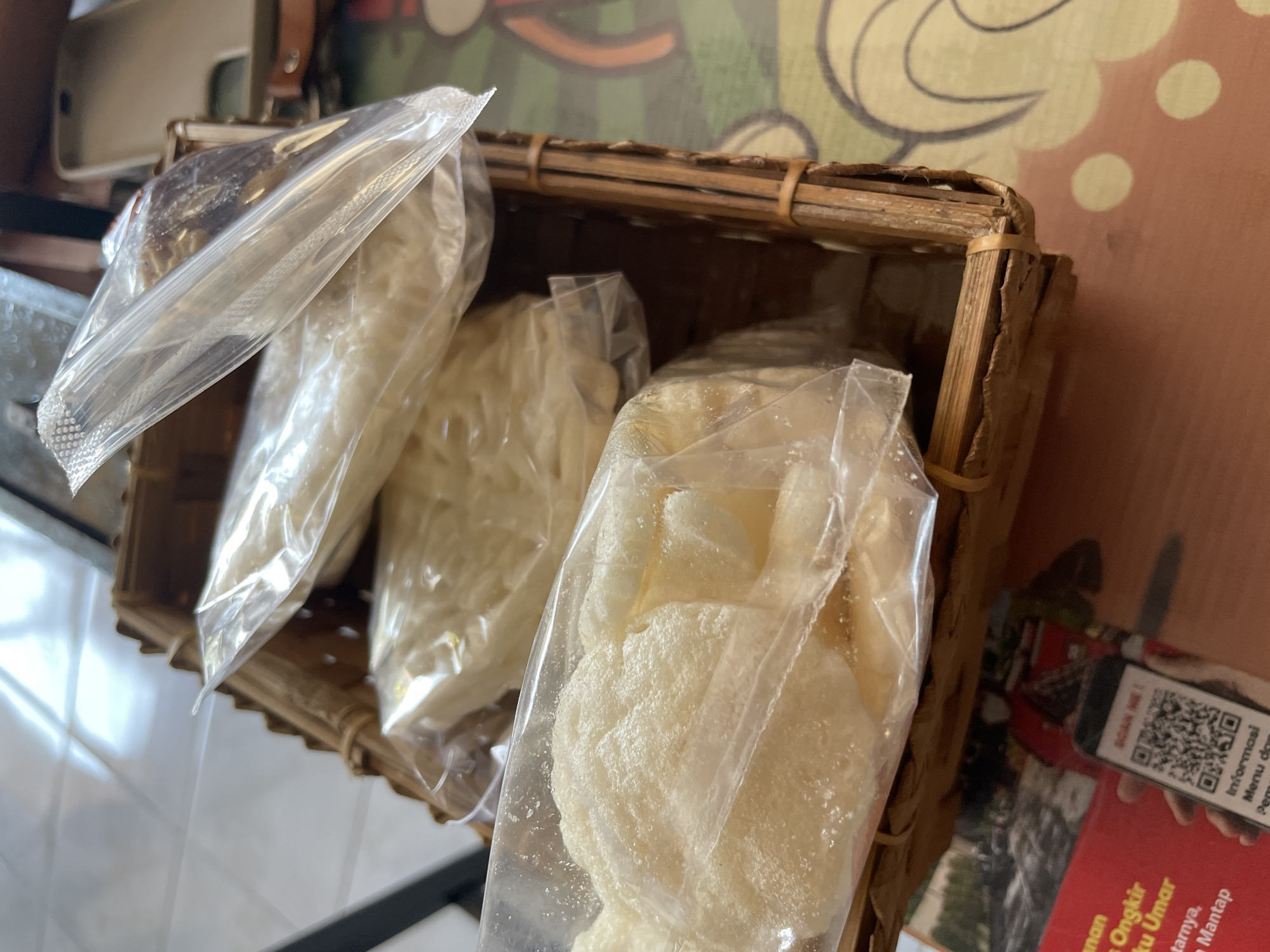 In Denpasar, Indonesia, this image depicts a close-up view of a woven bamboo basket filled with several plastic-wrapped servings of what appear to be puffed rice crackers or similar snacks. The basket is positioned on a table with a patterned surface. In the background, a light-colored object, possibly a mobile phone case, and part of a larger object, possibly a table or counter, are visible. The lighting suggests an indoor environment.