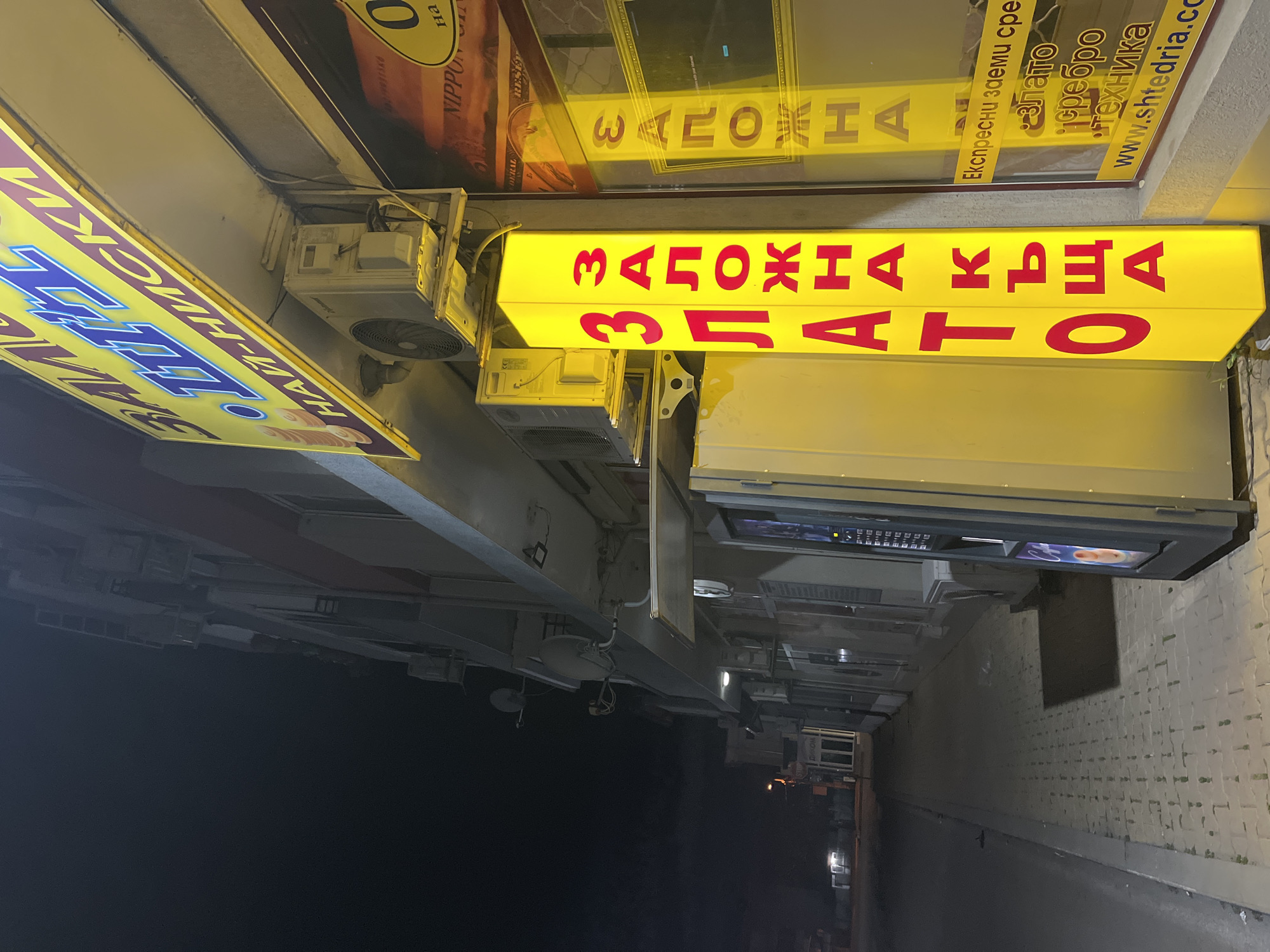 The image shows a nighttime street scene in Nova Zagora, Bulgaria, with illuminated signage in Cyrillic. The signs advertise "Заложна къща Злато," which translates to "Pawnshop Gold," along with information on express loans, gold, silver, electronics, and a website, www.shtedria.co. Additional signage includes what appears to be "BAKI" with smaller text below. An air conditioning unit is visible near the signs, and the background shows a narrow street with a brick wall on one side.