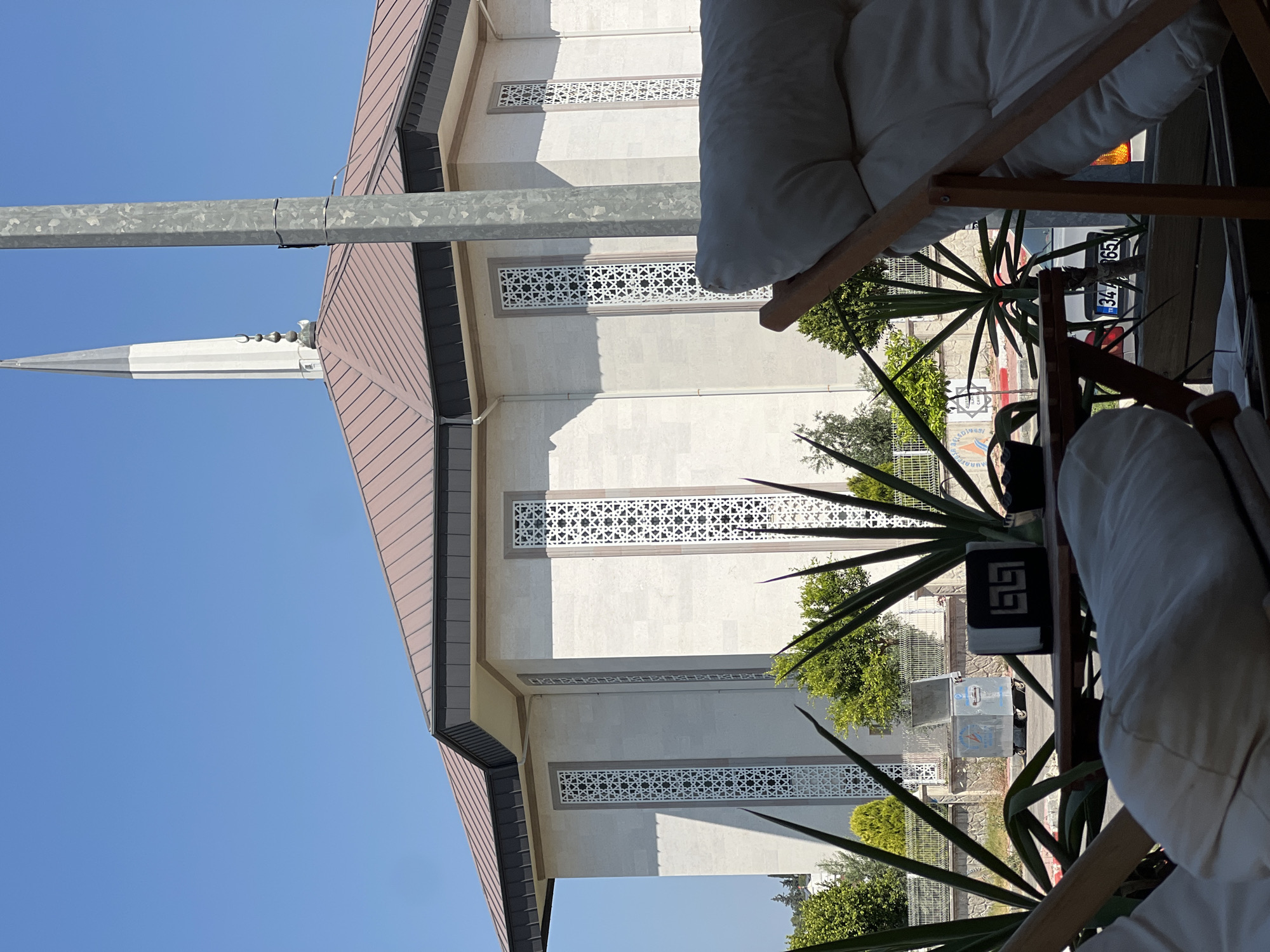 The image captures a view from a seating area, possibly a cafe or restaurant patio, looking towards a modern mosque. In the foreground, the corner of a light-colored armchair with a wooden frame is visible, along with a portion of another similar chair and a low wooden table. On the table, there's a black napkin holder with a geometric pattern and what appears to be a small dark object, perhaps a sugar packet or a spice holder.
Beyond the seating area, a large, light-colored mosque dominates the background. It features a large, light brown, sloped roof with dark grey trim. A tall, slender minaret with a pointed spire rises from behind the roofline. The mosque's exterior walls are constructed of light-colored stone blocks, and several tall, narrow windows with intricate white geometric lattice work are evenly spaced along the facade.
In the middle ground, between the foreground seating and the mosque, there's a chain-link fence and some green foliage, including spiky plants that resemble agave or yucca. A large, metallic dumpster with its lid open is visible behind the fence. In the far background, more trees and a clear blue sky complete the scene. A metal utility pole is positioned prominently on the right side of the image, partially obscuring the mosque. The overall impression is a sunny day in an urban setting, with a blend of modern architecture and greenery. The license plate visible on a car in the background, "34-xxx-65," suggests the location might be within Turkey, specifically Istanbul (indicated by the "34" plate prefix), though the prompt specifies Antalya, Turkey, where different plate prefixes are used.
