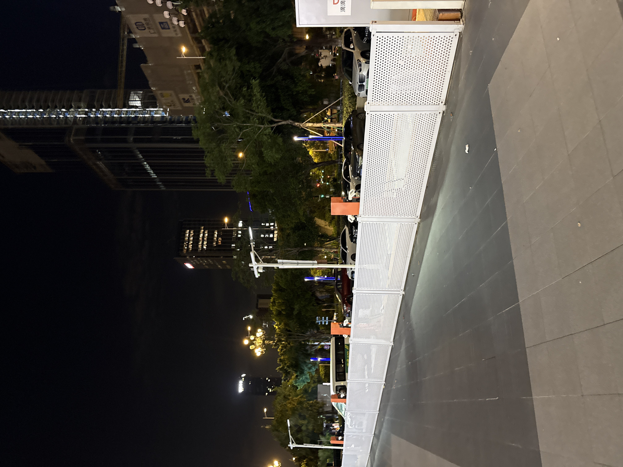 A nighttime, eye-level, medium shot depicts a street scene in Huangpu District, China. In the foreground, a grey tiled pathway transitions to a dark tiled surface, bordered by a white, perforated metal fence. Beyond the fence, a row of parked cars, including a white SUV and a black sedan, are visible. A white van is parked further down the street. Tall trees with green foliage line the street, interspersed with streetlights. In the background, illuminated by the night sky, are several high-rise buildings, some under construction, with lights shining from their windows. The overall lighting suggests an urban environment at night.
