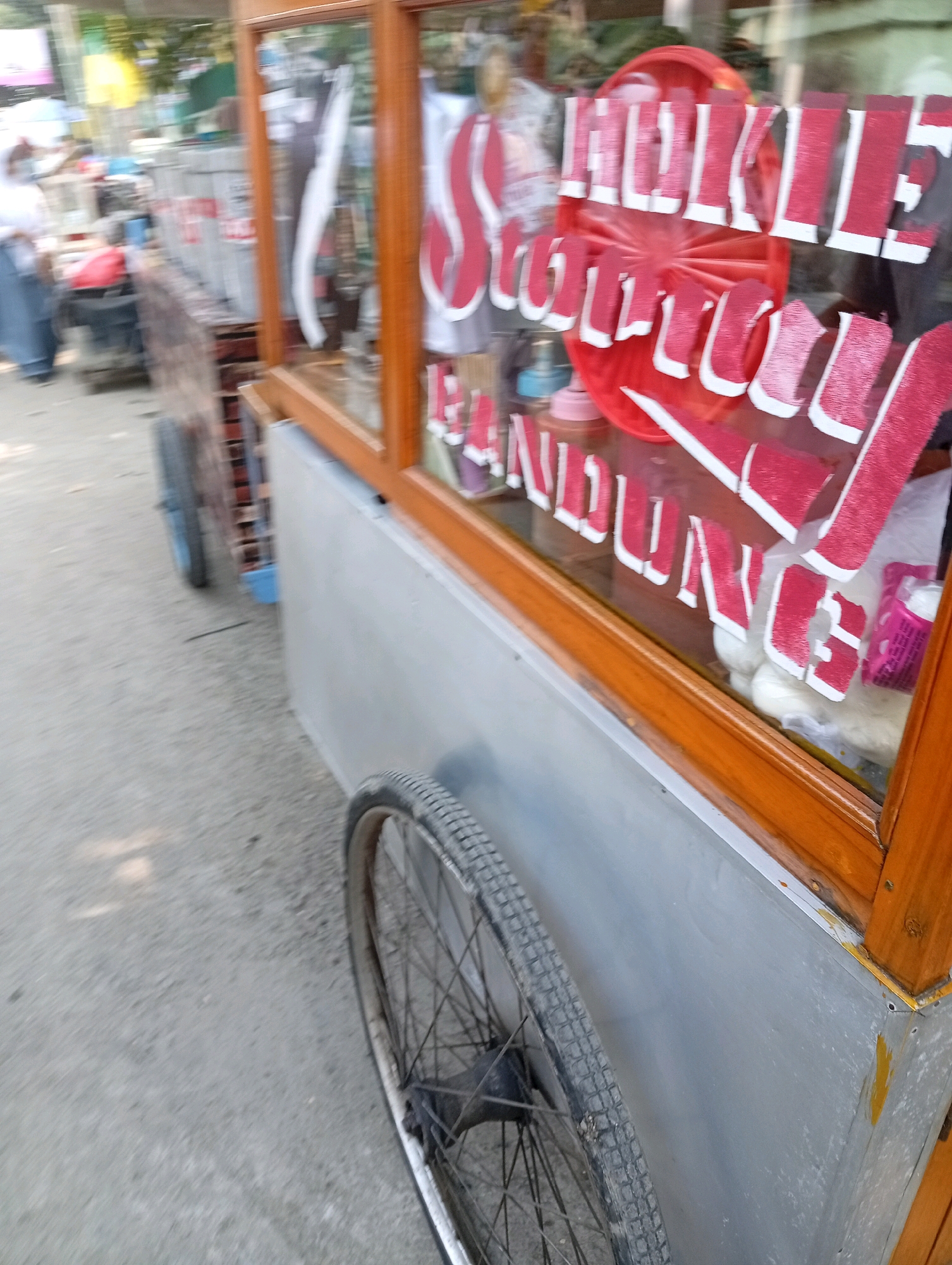 The image shows a close-up of a mobile food cart with red and white lettering on its glass window. The lettering appears to be in Indonesian, with some words partially obscured. The cart has a wooden frame around the glass and a metal body. A large bicycle wheel is visible on the left side of the cart, and another wheel is partially visible on the lower right. The cart is on a paved surface, possibly a street or sidewalk. In the background, there is a blurred view of a person and other objects, suggesting an outdoor setting. The overall impression is that the image was taken while the cart was in motion. The lettering on the cart suggests it might be a food vendor selling products originating from or related to Bandung, Indonesia, a city in West Java, but the question specifically mentions East Jakarta, Indonesia, which is a separate administrative city. Without further context, it's not possible to definitively place the cart in East Jakarta.