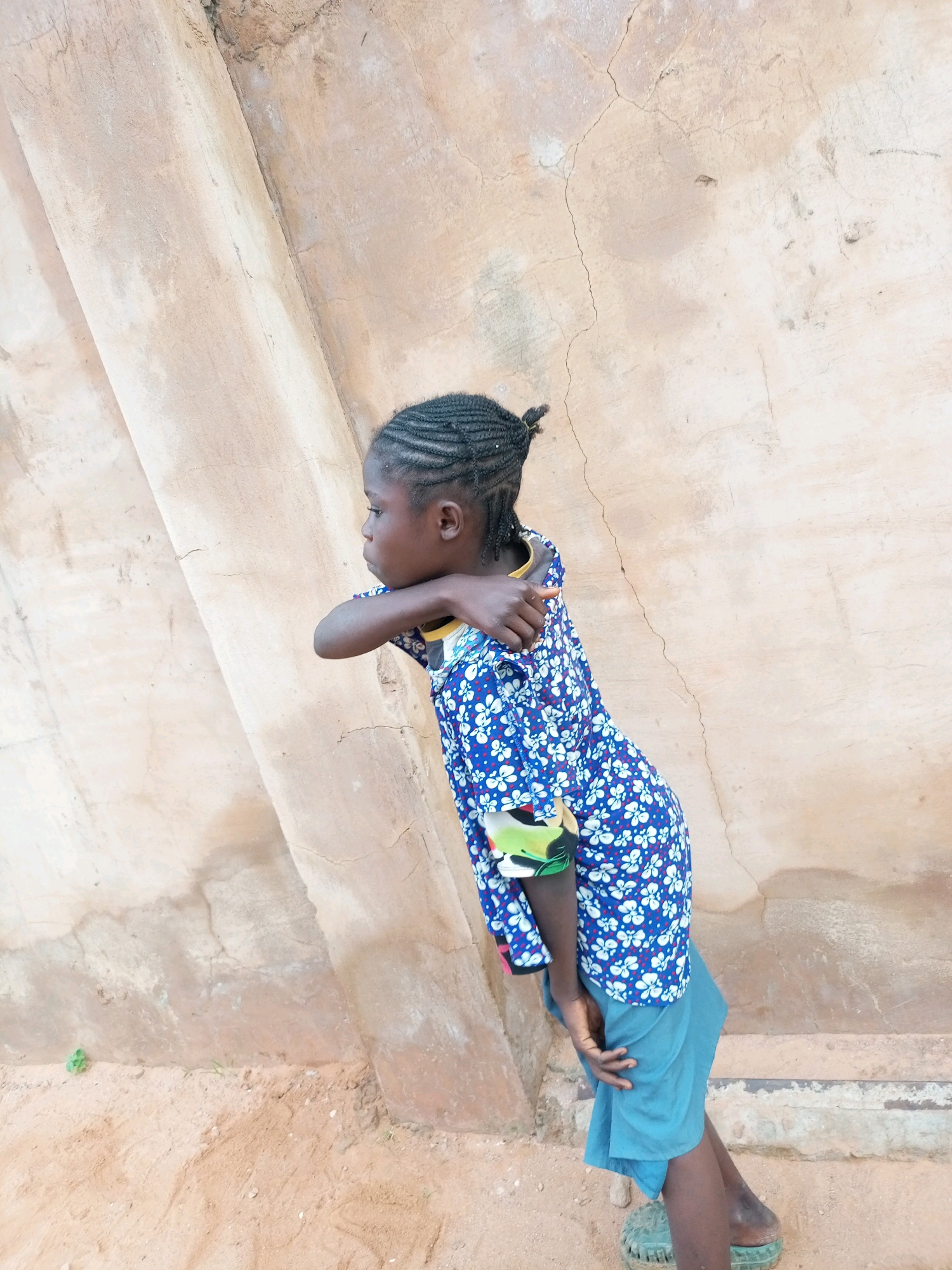 A young girl with braided hair stands against a textured, light-colored wall in what appears to be an outdoor setting in Tempure, Nigeria. She is wearing a blue, floral-patterned shirt with white flowers and a light blue skirt. Her left arm is bent and resting on her chest, with her hand near her shoulder. Her right arm is behind her, with her hand visible near her hip. She is looking to her left, away from the camera. The ground in front of her is sandy and appears to have some small green plants growing. The wall behind her is cracked and shows variations in color, suggesting age and exposure to the elements. The scene is illuminated by natural light, casting subtle shadows.