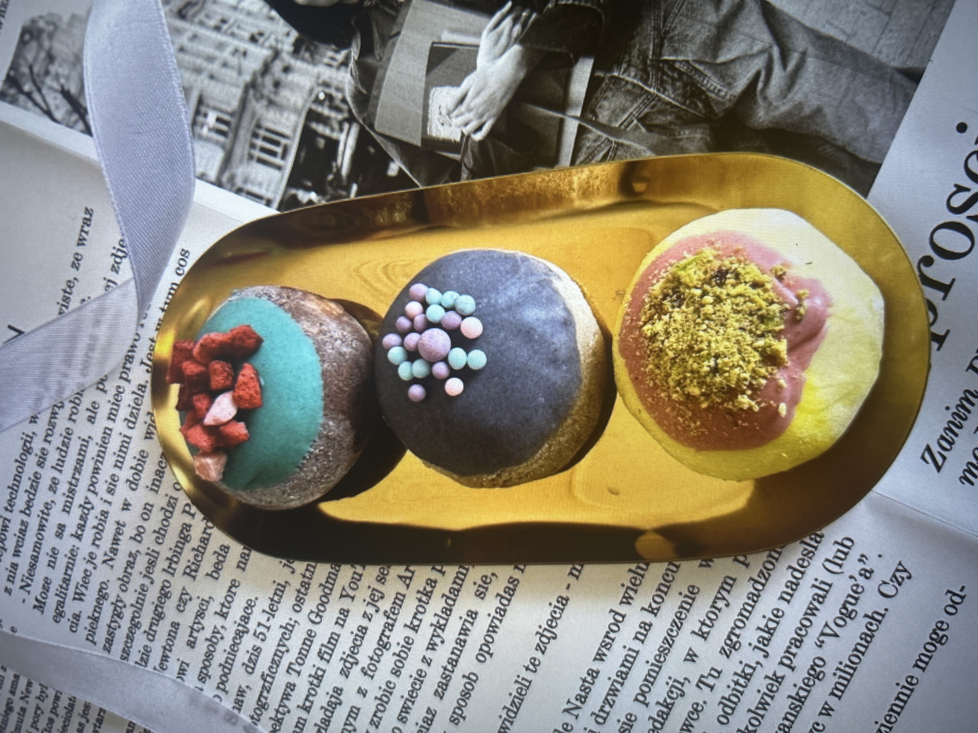 The image shows three gourmet doughnuts arranged on a rectangular, gold-colored tray. The doughnut on the left has a turquoise frosting and is decorated with small red pieces. The center doughnut features a dark gray frosting and is adorned with multicolored pastel-colored balls. The doughnut on the right has a yellow base with a pink swirl and is topped with finely chopped green pistachios.
The tray sits on top of a newspaper with Polish text. The background is partially obscured, showing a black and white photograph of hands and a light gray ribbon.
There is no information related to the city of Câmara de Lobos, Portugal, present in the image.