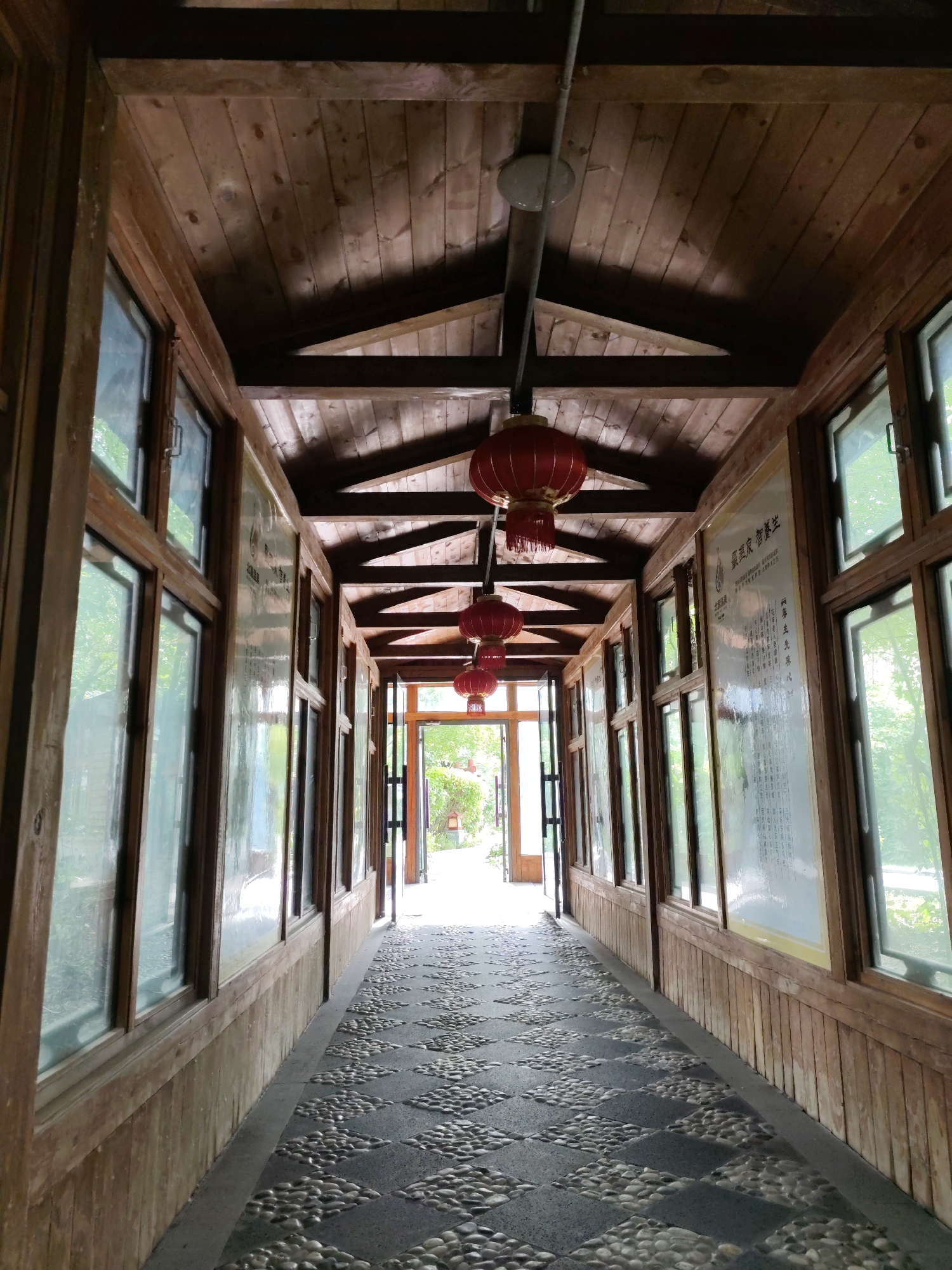 The image depicts a long, covered wooden walkway with a pebble and tile floor. The walkway is lined with windows on both sides, and red Chinese lanterns hang from the ceiling. At the far end of the walkway, double doors lead outside to a bright, green area with trees. The scene appears to be in a park or garden setting within Lindian, China. There are no people visible in the image. The wooden structure shows a rustic, natural aesthetic, and the lighting suggests it is daytime.