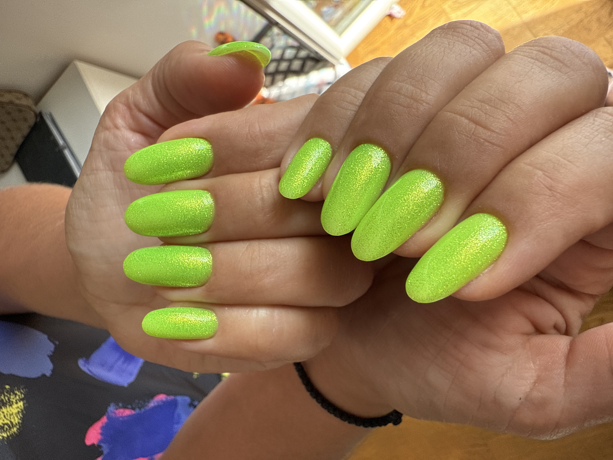 A close-up shot shows a person's hands with bright neon green, glitter-infused oval-shaped nails. The hands are positioned with fingers slightly curled and overlapping, displaying ten nails in total. The nails are well-maintained and uniformly shaped. On the left wrist, a thin black braided bracelet is visible. The background is softly blurred, with hints of colorful abstract patterns on clothing in the upper left corner and a light-colored surface with a patterned object and a mirror or reflective surface in the upper right background. The lighting suggests an indoor setting with warm ambient light.