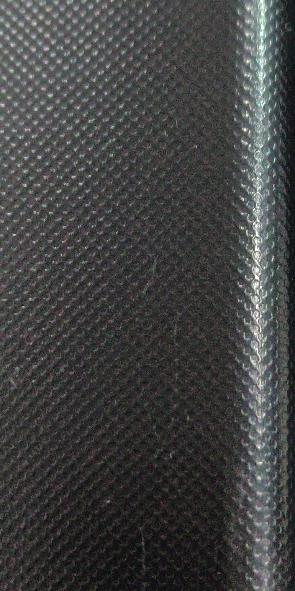 The image is a close-up, abstract shot of a textured dark surface, possibly fabric or rubber, with a vertical metallic-looking strip on the right side. The dark surface has a repetitive pattern of small, raised dots or bumps, creating a tactile appearance. Light reflects off these bumps, giving them a subtle shimmer. The metallic strip on the right exhibits a more pronounced textured pattern, appearing to be made of interconnected, curved elements that catch the light with a silvery sheen. The overall impression is one of a material with a robust and possibly protective quality, due to the textured and possibly durable materials depicted.