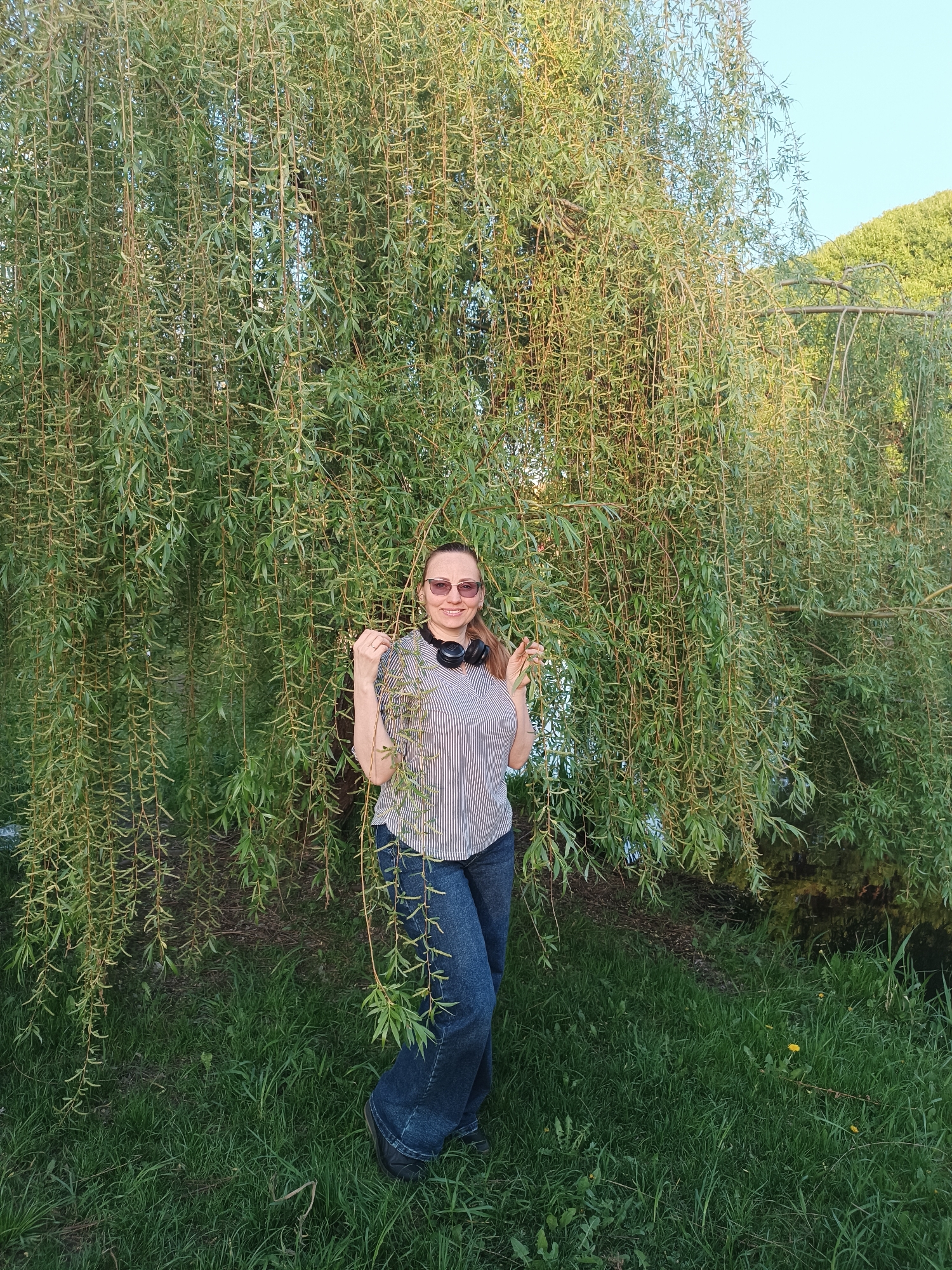 A medium shot captures a woman standing in front of a large weeping willow tree. She is wearing sunglasses, a striped shirt, and jeans, with headphones around her neck. The background consists of a grassy area with a hint of a body of water and additional foliage, suggesting an outdoor setting. The location is the city of Unknown, Unknown.