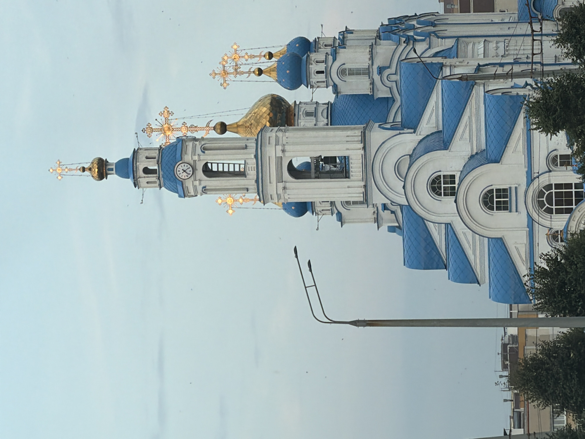 A white and blue church with golden domes and crosses stands under a cloudy sky. A street light pole is visible in the foreground to the left of the church. The church features multiple blue, arched roofs and ornate white architectural details. Some smaller buildings are visible in the background behind the church, partially obscured by trees. The sky is a pale blue with some wispy clouds.