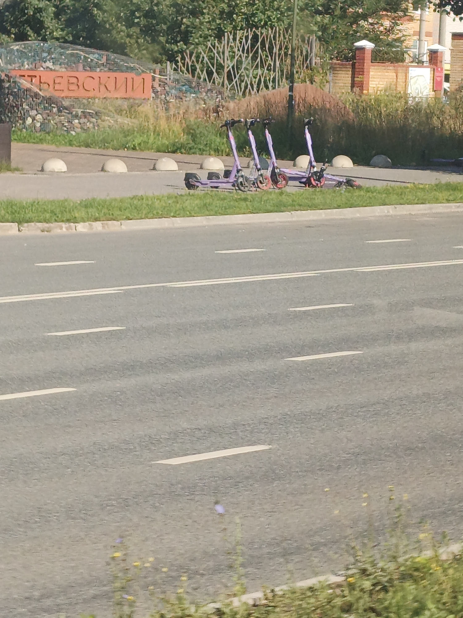 The image depicts an outdoor scene with a street in the foreground and a park or public space in the background. A row of purple electric scooters is parked on the sidewalk near a sign with Russian lettering. The sign appears to say "НЕВСКИИ" which translates to "Nevsky" or "Nevsky Prospekt," a famous street in Saint Petersburg, Russia. However, the user has specified that the location is Kazan, Russia. The background shows trees, a fence, and brick buildings. The street has lane markings. There are no people visible in the image.