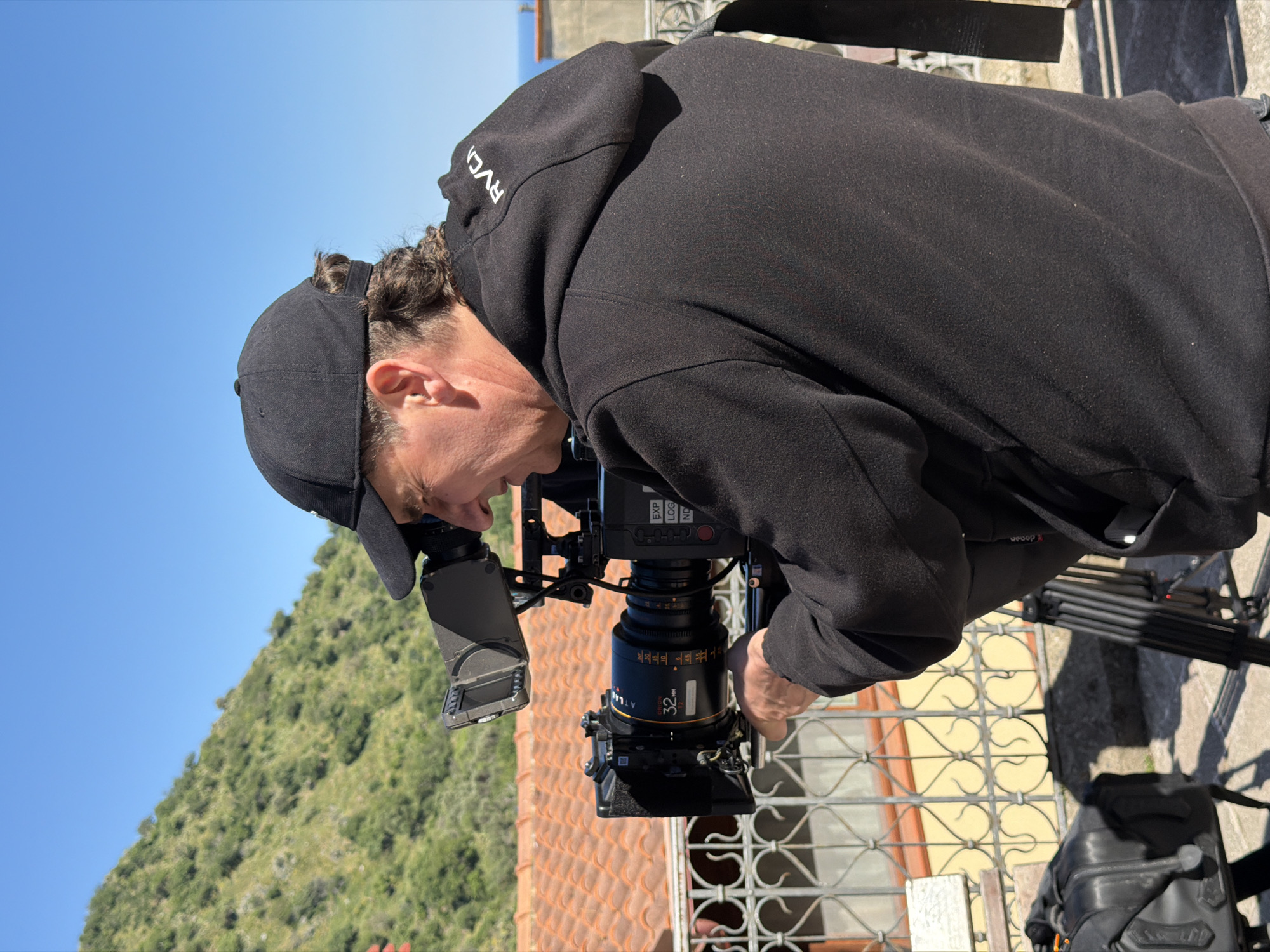 A man, seen in profile, is intently operating a professional video camera outdoors under a clear, bright blue sky. He wears a black baseball cap and a black hooded sweatshirt with "RVCA" visible in white lettering on the hood. His dark hair is pulled back.
The man is focused on the camera setup, possibly looking through a viewfinder or at an attached monitor. The camera is a substantial piece of equipment with a large lens clearly marked "ATLAS" and "32mm T2.0". Text such as "EXP" and "LOG" are also visible on the camera body. The camera is mounted on a tripod, part of which is visible to the right.
In the background, a verdant, bushy mountain rises against the sky on the left. In the mid-ground, a terracotta-tiled roof and an ornate, light-colored metal railing with a decorative, looping pattern are visible, suggesting an architectural setting. The lighting indicates a sunny day. The scene captures a professional filmmaking activity in Itala, Italy.