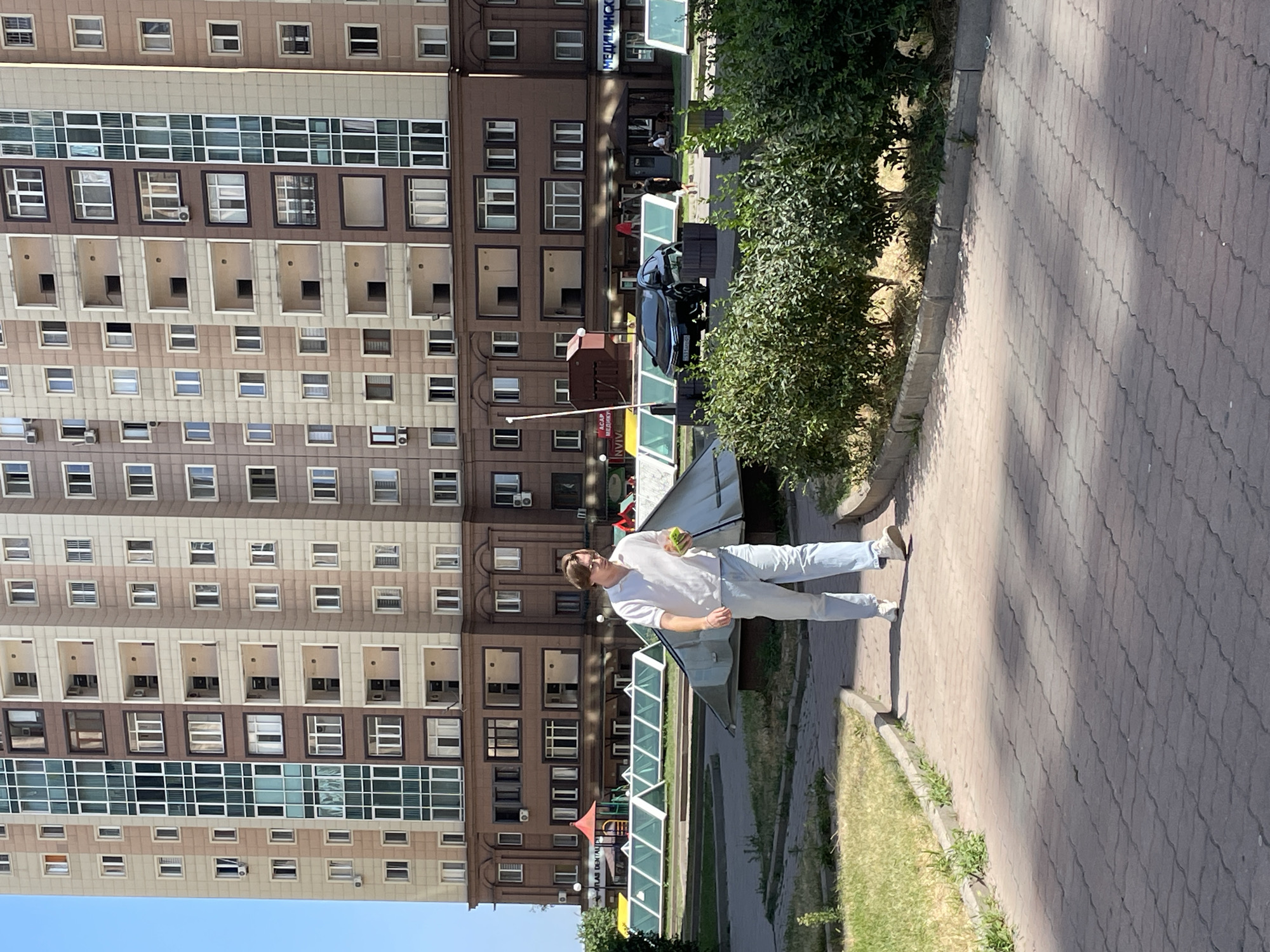 A young man with blond hair, wearing a white polo shirt and light blue jeans, walks along a paved walkway. He is holding a small green object in his right hand. In the background, a large, multi-story apartment building with beige and brown facades and numerous windows stands under a clear blue sky. To the left of the walkway, a patch of grass is visible. Further in the background, a black car is parked. The environment suggests a residential area within the city of Almaty, Kazakhstan, with the architectural style and the presence of greenery indicating a developed urban setting.