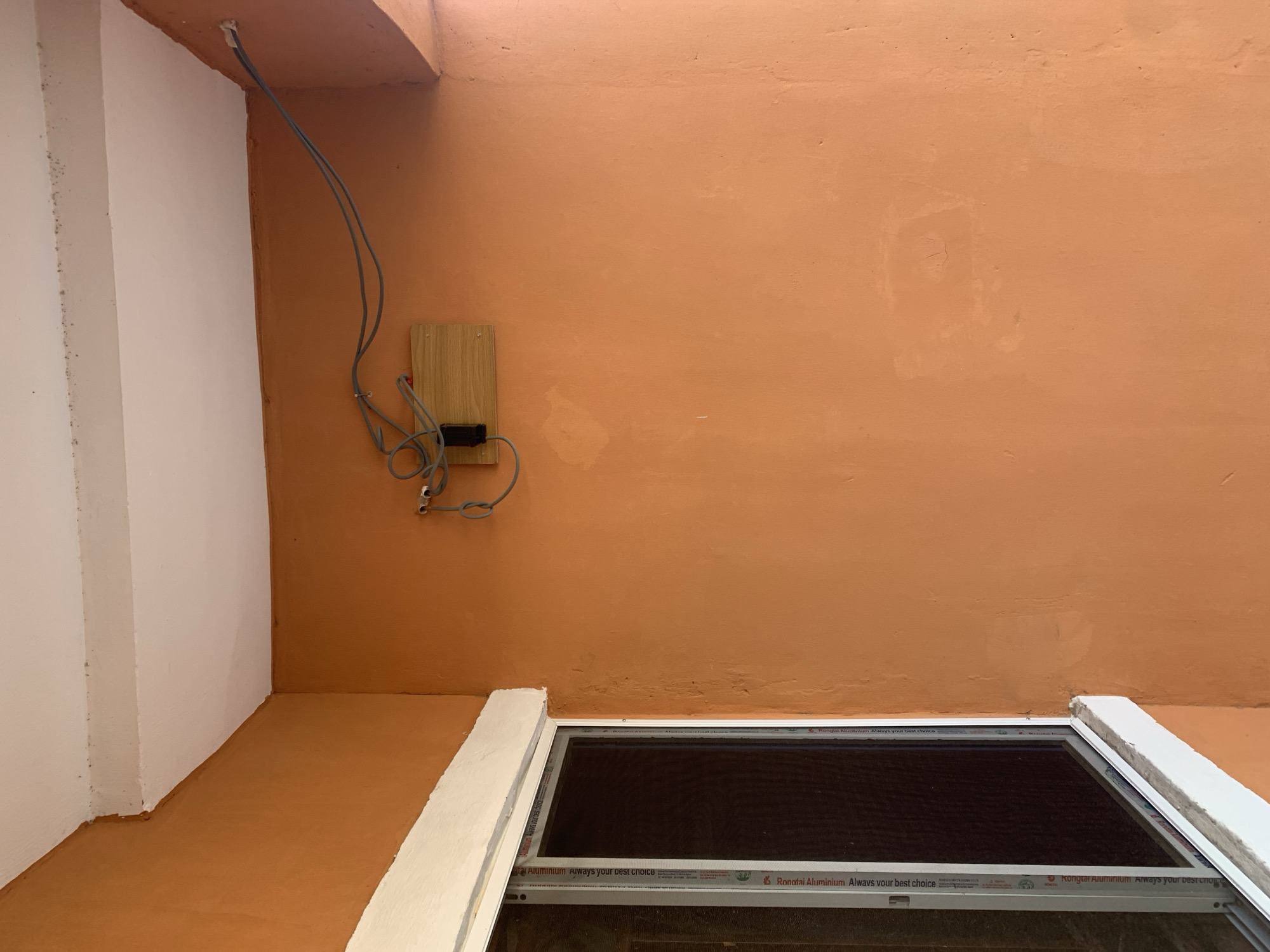 An outdoor corner of a building features an orange-painted wall and a white ceiling. A window with a dark screen is visible on the left side. Mounted on the wall is a small wooden electrical box with gray wires connected to it, extending upwards to the ceiling. The overall scene is simple and functional.
