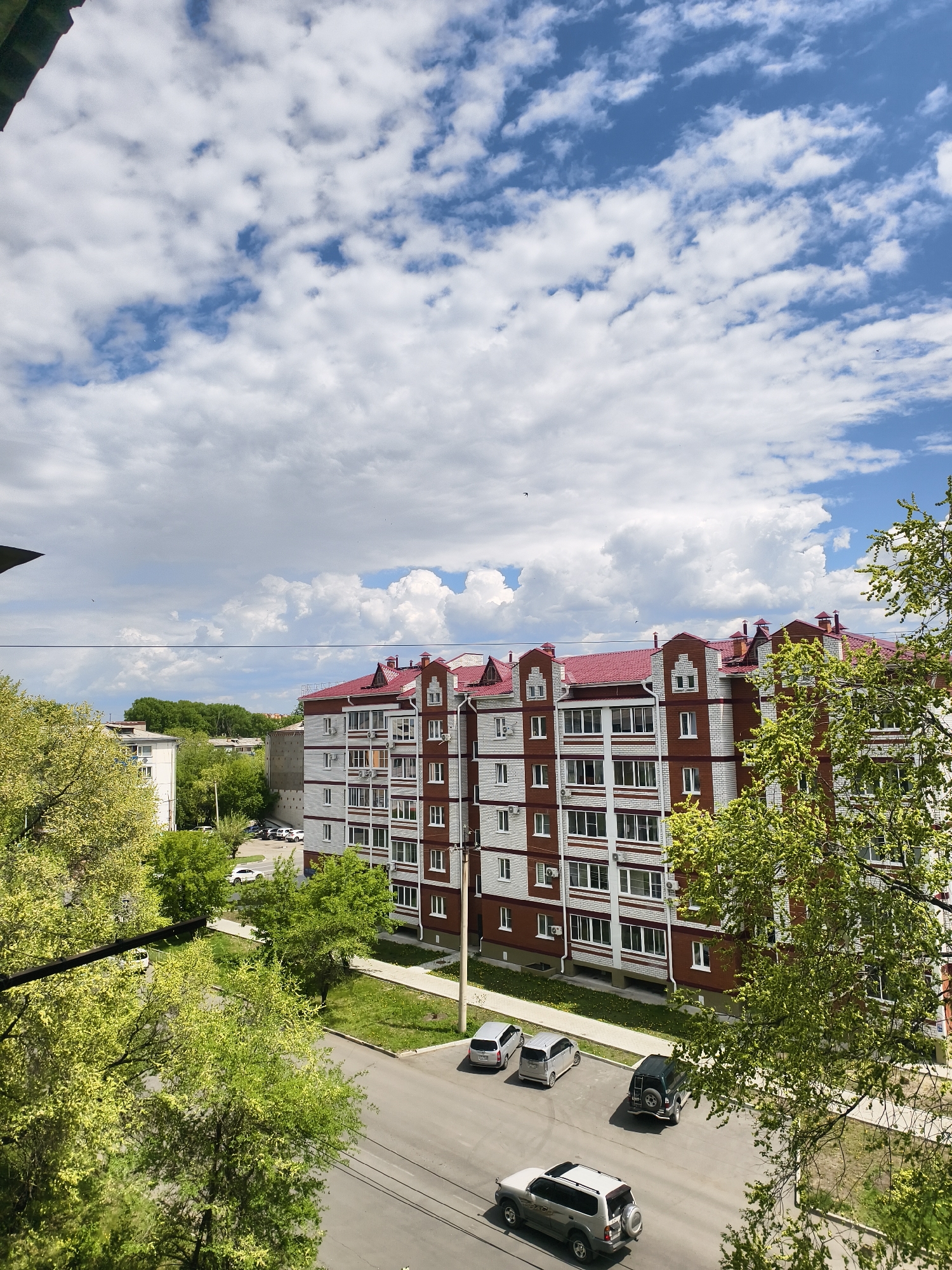 The image shows a view from a high vantage point overlooking a residential street in Belogorsk, Russia. The foreground is dominated by green trees with yellow blossoms and branches. In the middle ground, a red-roofed, multi-story apartment building with white and brown paneling is prominently featured. A few cars are parked along the street in front of the building. The street itself is asphalt and shows signs of wear.
Beyond the buildings and trees in the background, a strip of green trees is visible along the horizon. The sky takes up a significant portion of the frame, filled with fluffy white clouds scattered against a blue background. The overall lighting is bright, indicating a sunny day. The view is typical of a residential area in Belogorsk.