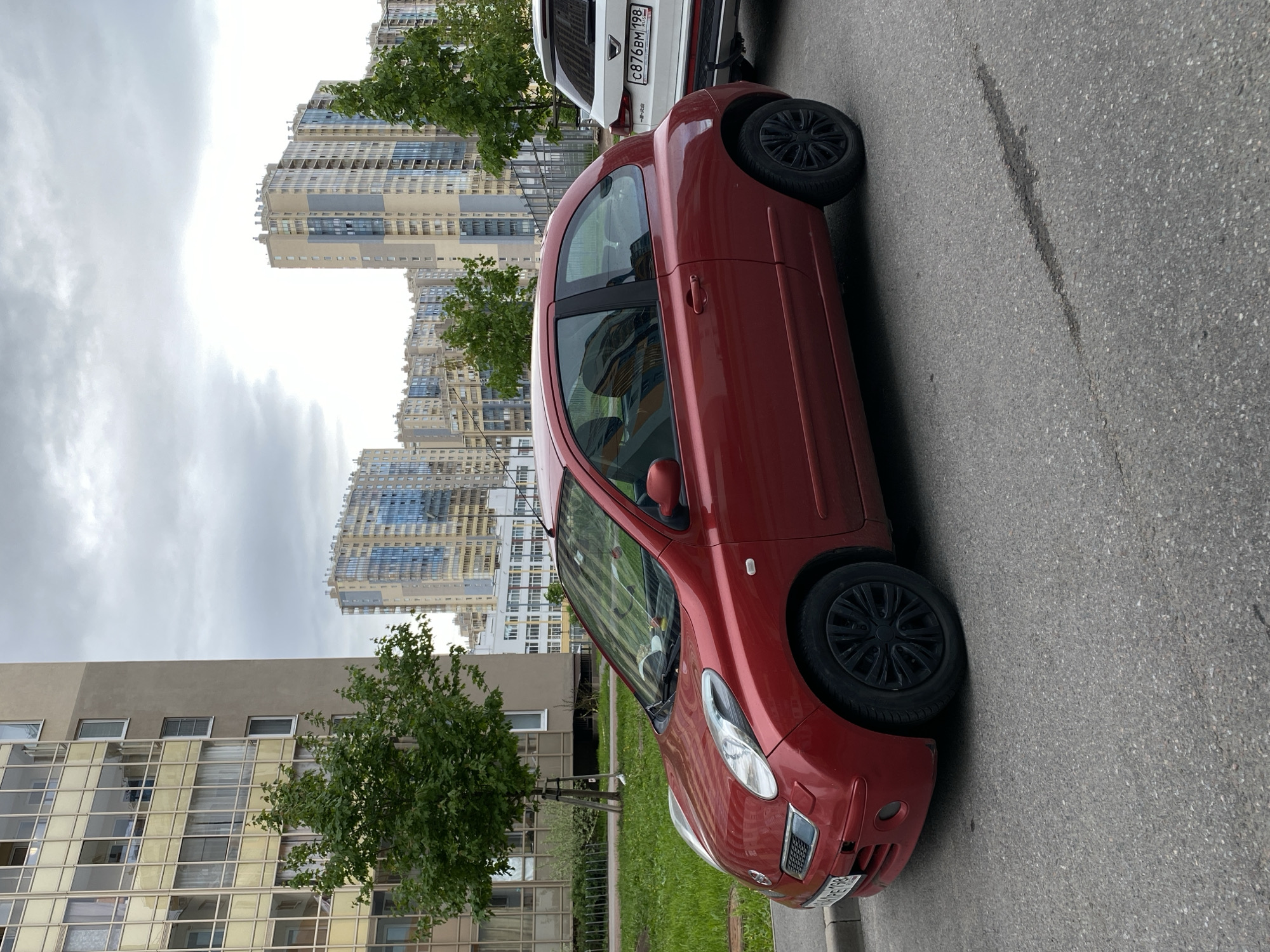 A red Nissan Micra is parked on the side of a street in Saint Petersburg, Russia. The car is situated next to a curb with a small patch of grass and a young tree. In the background, there are apartment buildings, some of which are tall and modern with many windows. The sky is overcast. The license plate of the car is partially visible. Another car, white, is parked just behind the red Nissan.