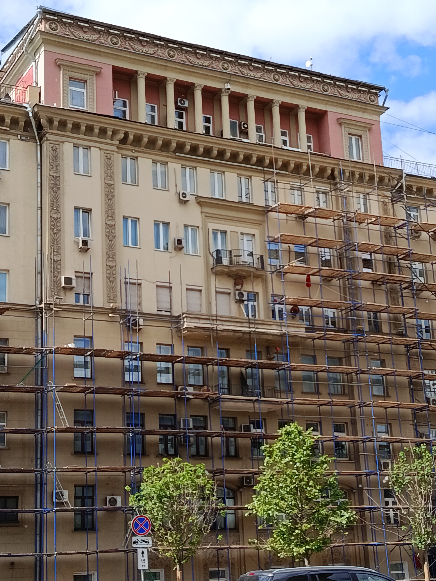 An outdoor, eye-level, full shot captures a multi-story building in Moscow, Russia, largely obscured by extensive scaffolding, suggesting ongoing renovation or maintenance work. The architecture is grand, reminiscent of neoclassical or Stalinist Empire style, featuring ornate details.
The uppermost floor stands out with its distinct pink or salmon color, featuring a prominent colonnade of several tall, light-colored columns supporting an elaborate, intricately patterned cornice. This floor has multiple windows, some with air conditioning units installed below them. The roofline above displays decorative elements, including star-like motifs.
Below this, the main facade of the building is beige, adorned with decorative pilasters and cornices. Numerous windows are visible across its surface, many with exterior air conditioning units attached. A substantial metal scaffolding structure, characterized by blue vertical poles and wooden platforms, covers the majority of the building's lower and mid-sections, allowing glimpses of the windows through its framework. Ladders and what appear to be work lights are visible on the scaffolding.
At the bottom of the frame, a dark car, possibly a sedan or SUV, is partially visible. Two green trees with dense foliage are in the foreground, partially obscuring the lower levels of the building and the scaffolding.
A circular "No Stopping" traffic sign (a red cross on a blue background) is visible in the lower-middle foreground, mounted on a pole. Below it, a supplementary rectangular sign indicates "15 M" (15 meters) with an upward arrow, specifying the length and direction of the parking restriction. The sky above is blue with scattered white clouds, indicating a clear daytime setting.