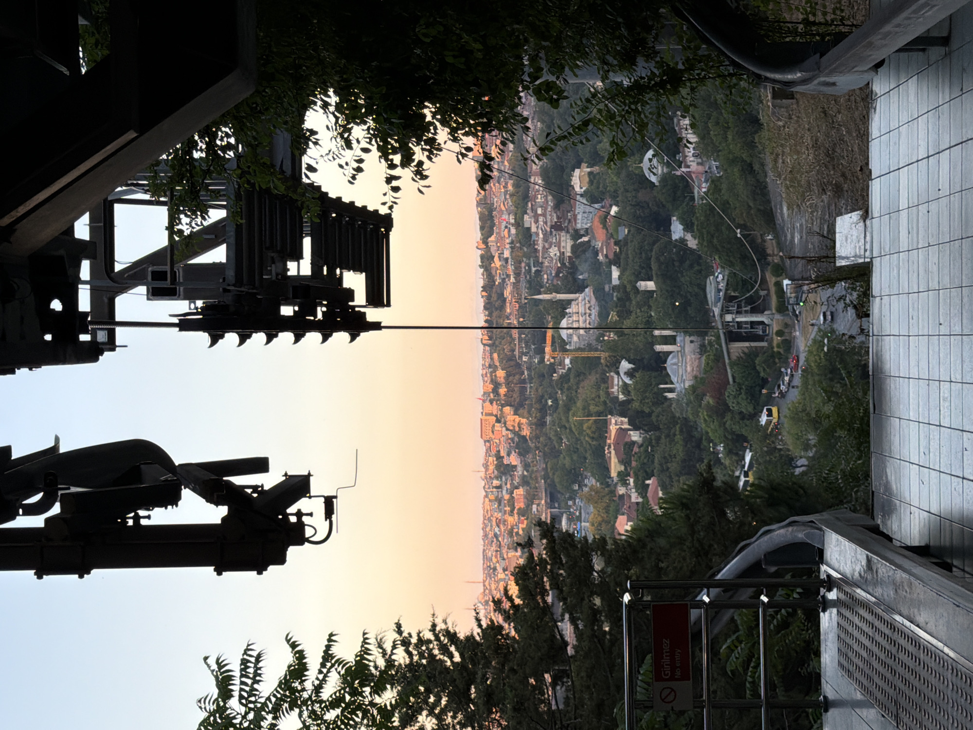 The photograph captures a view of Eyüpsultan, Turkey, from a high vantage point, likely a cable car station, during what appears to be sunset. The foreground is dominated by the dark, industrial structure of the cable car mechanism, silhouetted against a soft, orange and pink sky. Below the mechanism, a metal railing with a red "Girilmez No entry" sign is visible on the left. The platform in the immediate foreground is tiled with light-colored squares.
Beyond the station, the cityscape of Eyüpsultan unfolds. Numerous buildings with red-tiled roofs are densely packed, interspersed with lush green trees. Several mosques with distinctive domes and minarets punctuate the urban landscape, including what appears to be the Eyüp Sultan Mosque complex. A yellow bus and several cars are visible on a road winding through the city. The overall impression is of a historic city bathed in the warm glow of the setting sun.