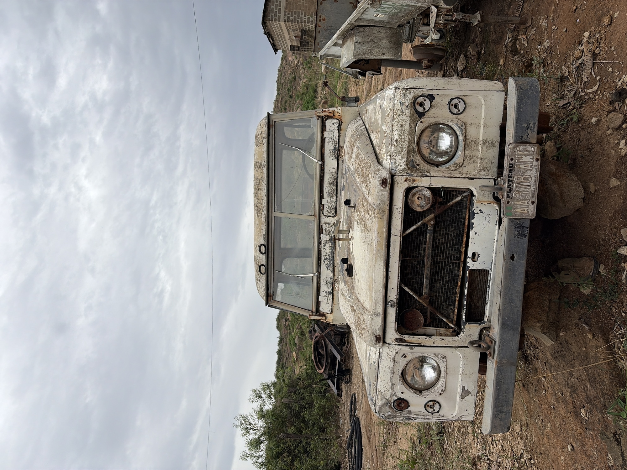 The image depicts a severely weathered white Land Rover abandoned in a dirt clearing. The vehicle's paint is heavily chipped and rusted. Its front grille is damaged. A license plate is partially obscured.
The background features a cloudy sky, low vegetation, and a dilapidated structure, possibly a building or shed.
The location is Gura Top, Nigeria. There are no people visible. The scene appears to be a vehicle junkyard.