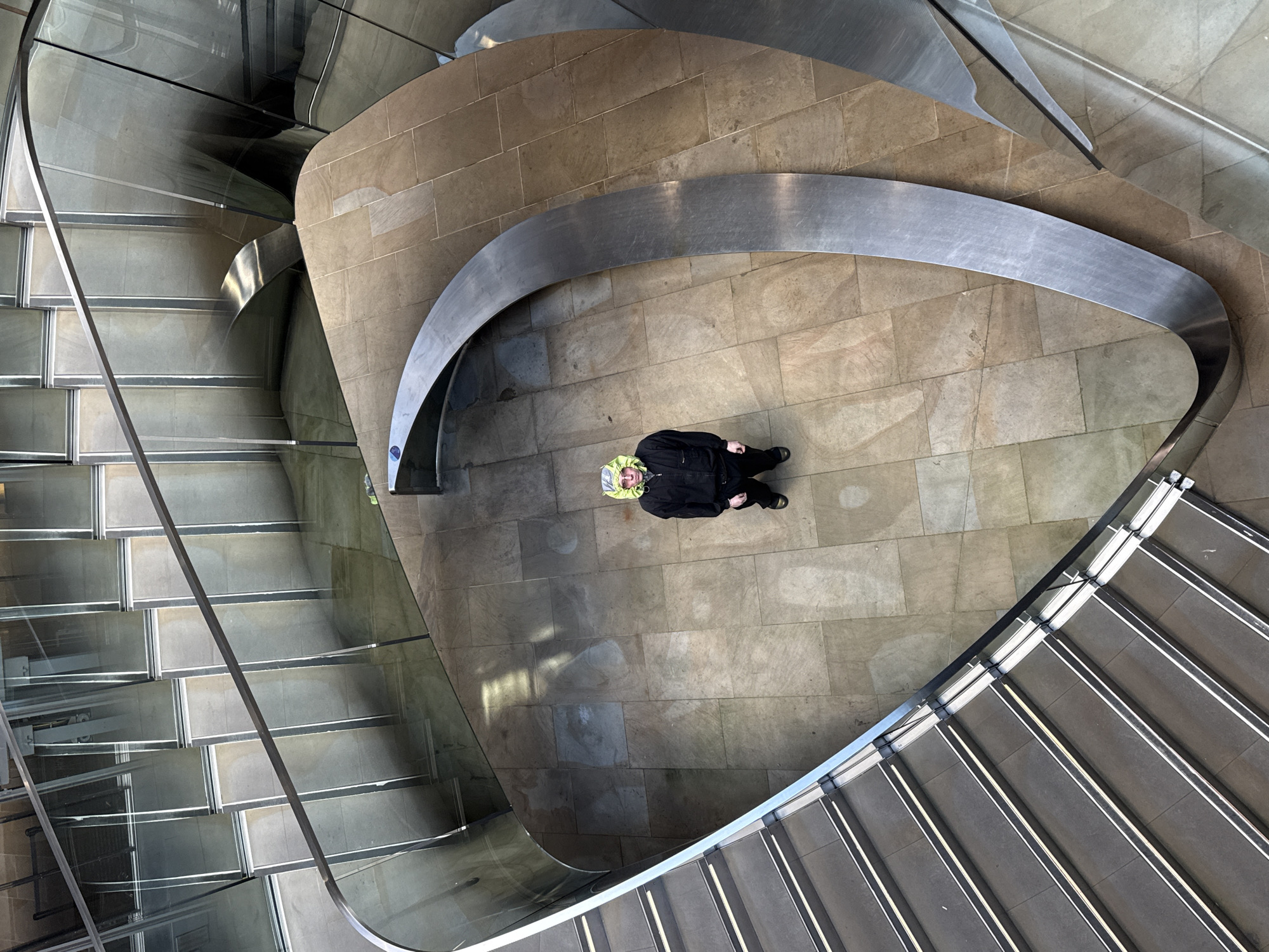 An elevated, top-down view depicts a person standing within a multi-story stairwell. The stairwell is composed of stone tiling, with staircases on either side of a central open area. Metal guardrails and glass enclosures are integrated into the staircase design. The person is wearing a black jacket and stands near a curved, decorative metal accent. The location is in London, United Kingdom.