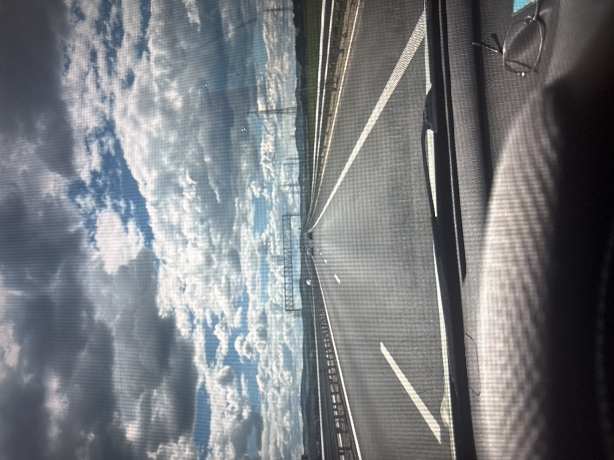 The image is a first-person perspective of a driver looking forward on a highway. The road is asphalt with white lane markings, and there are guardrails on the sides. In the distance, there are hills and power lines. The sky is filled with dramatic, dark clouds mixed with patches of blue sky. In the foreground, the dashboard of a car is visible, with a steering wheel on the left and a pair of glasses on the right. There are no people or specific landmarks that would indicate the location in Sofia, Bulgaria. The scene is consistent with driving on a major highway.